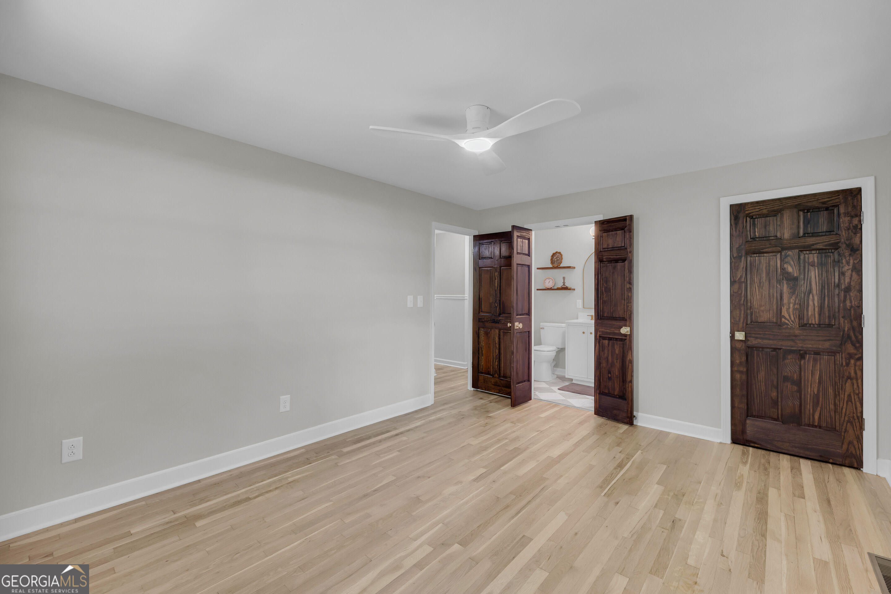 Vacant room with light wood floors, a ceiling fan, and open dark wood doors to a bathroom.