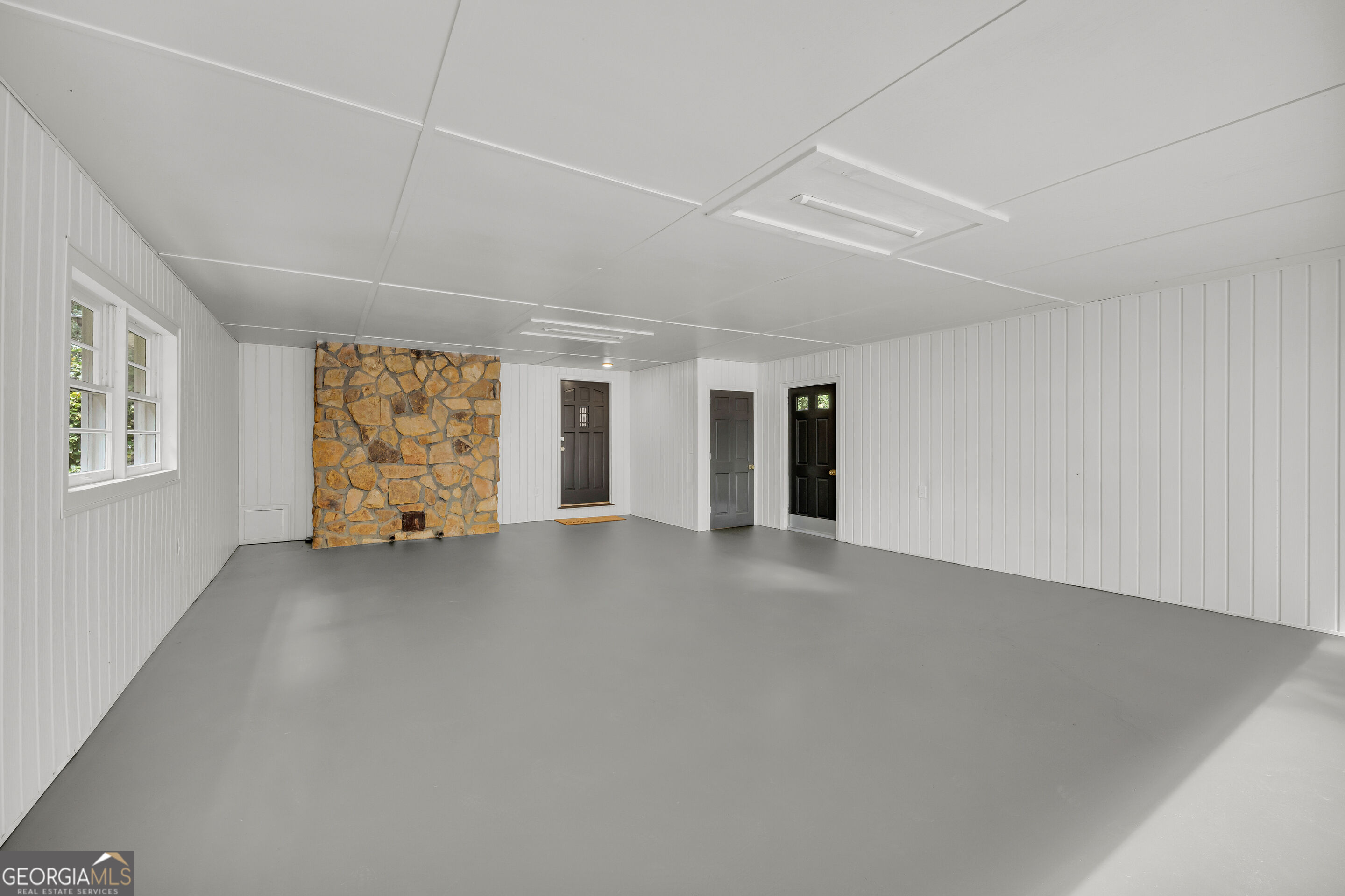 Vacant room with a stone fireplace, gray floor, white paneled walls, and several dark doors.