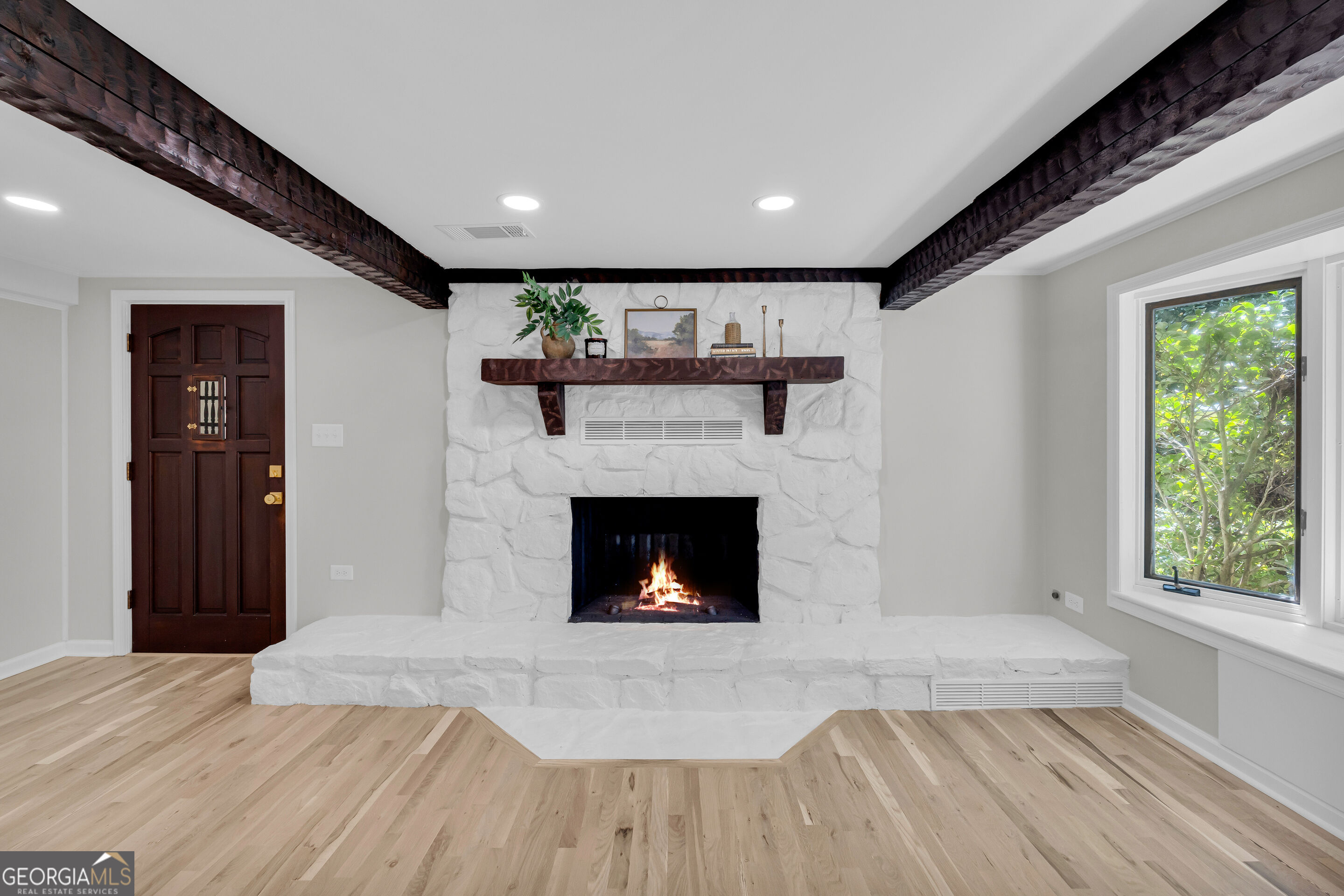 Living room with a white stone fireplace, lit fire, dark wood beams, and pale wood floors.