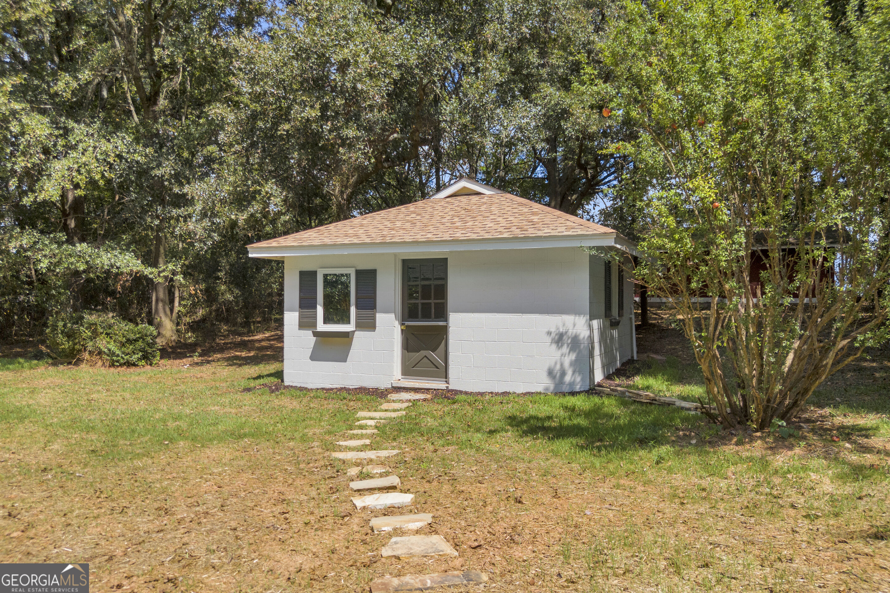 Small white outbuilding with a tan roof and stepping-stone path in a wooded yard.