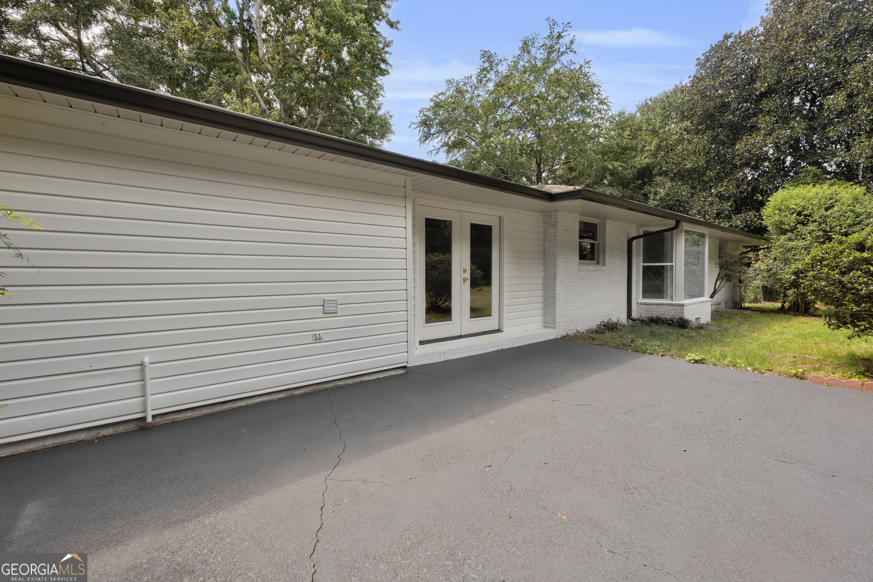 Single-story white house with double entry doors and a broad paved driveway, surrounded by mature trees.