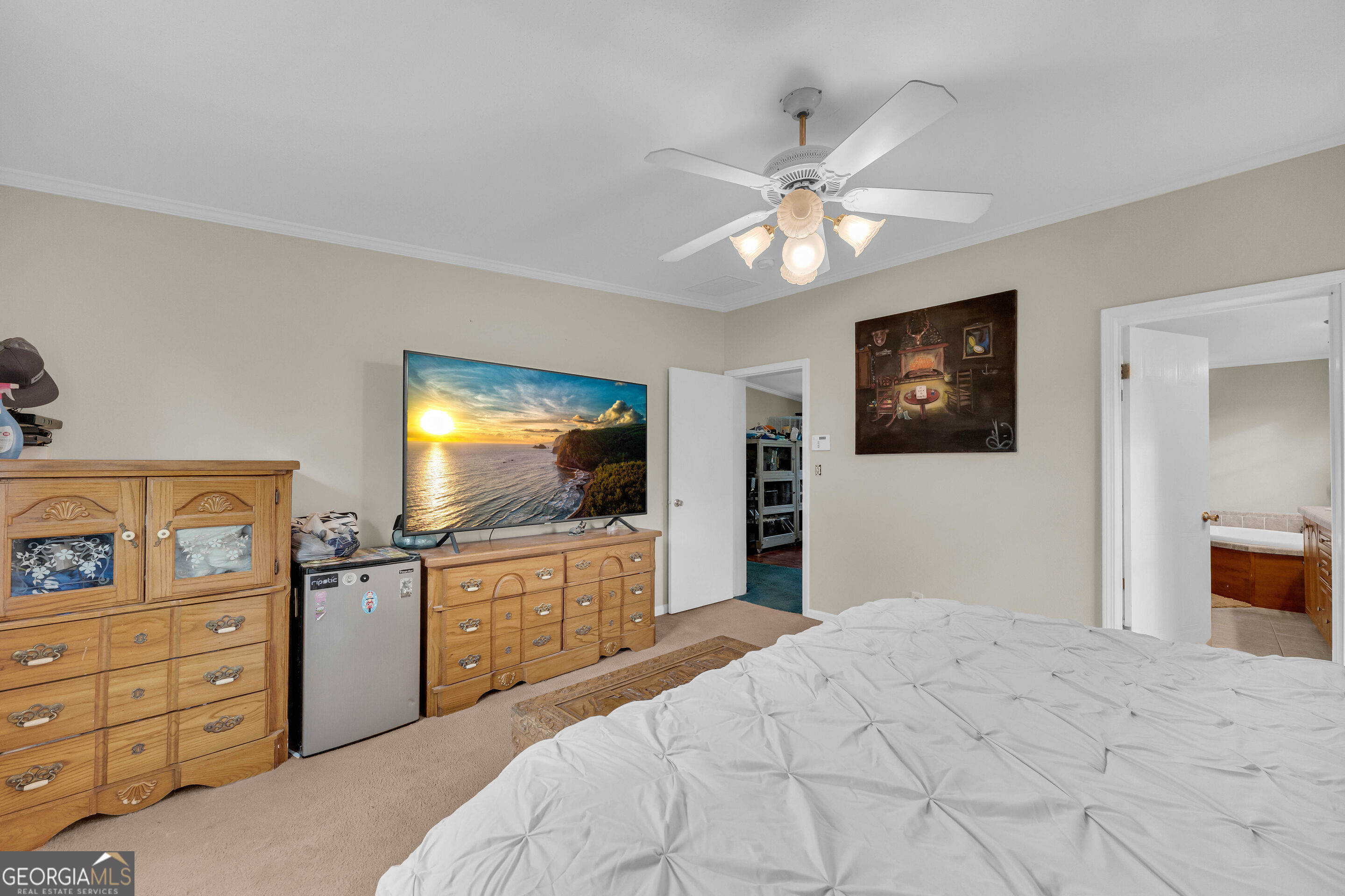 Furnished bedroom with a white bed, wooden dressers, and a television showing a coastal sunset.