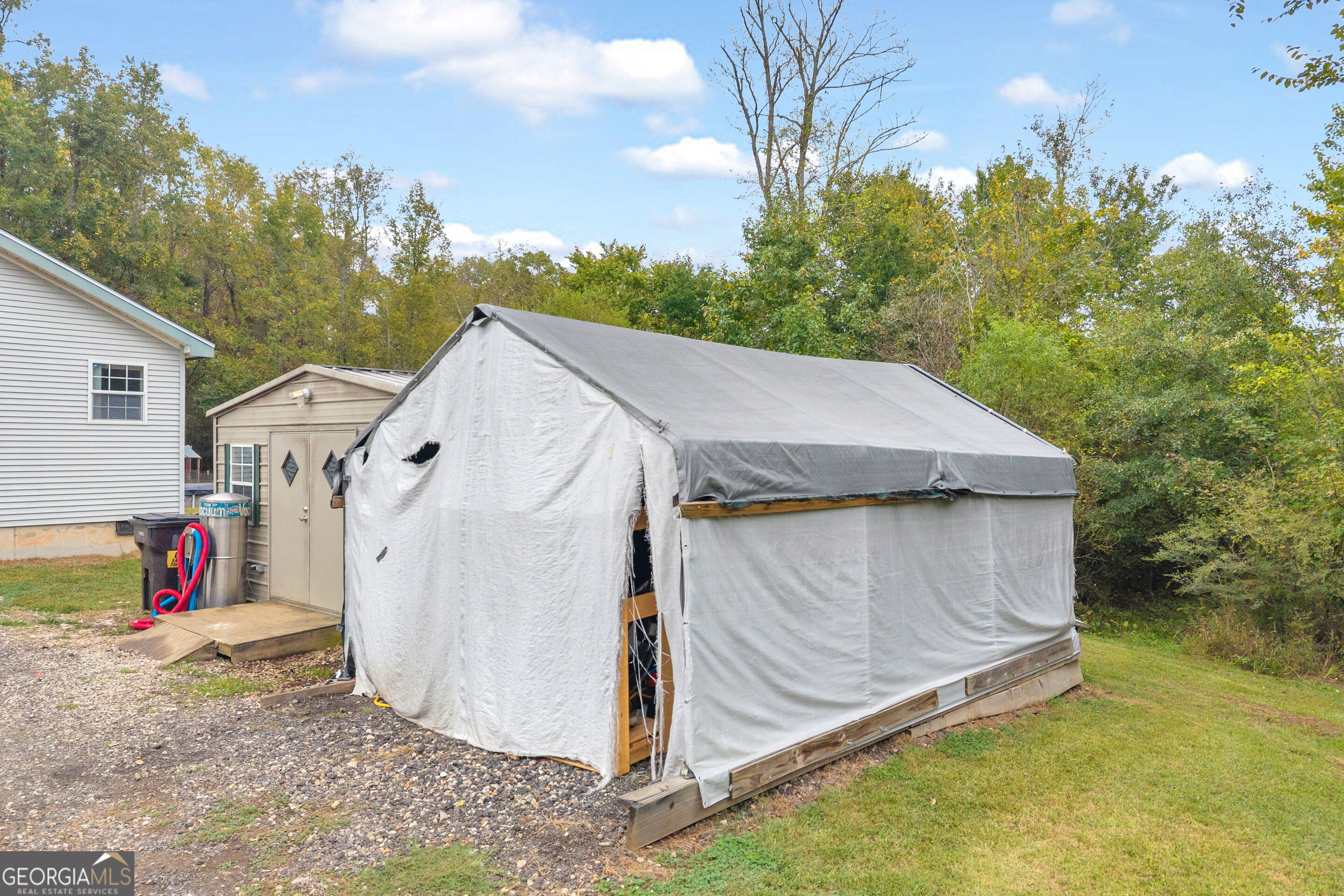 Tarp-covered outbuilding beside a small shed, with a house and wooded yard behind it.