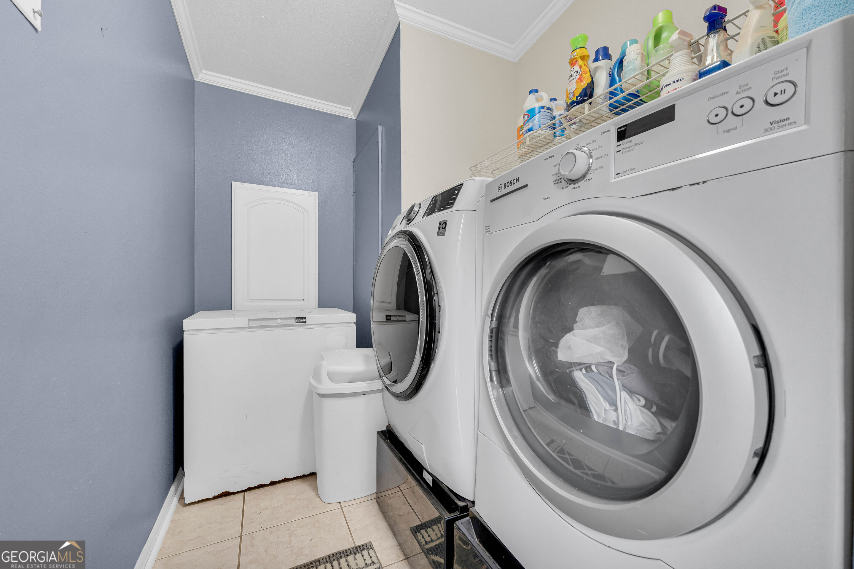 Laundry room with front-loading washer and dryer, top-loading washer, and cleaning supplies on a shelf.
