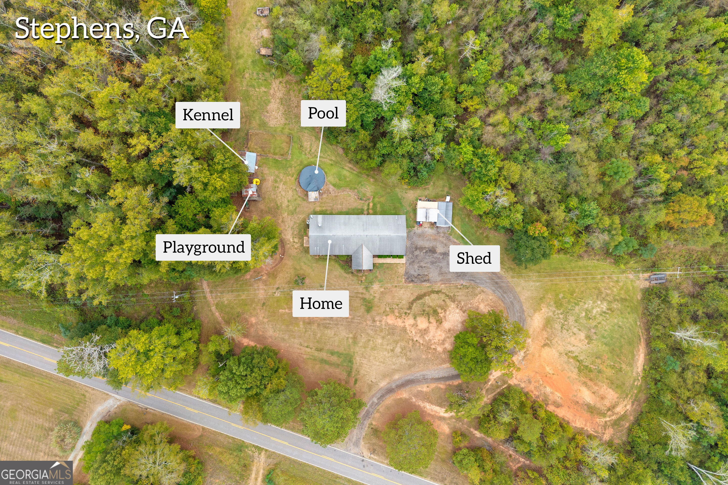 Aerial view of a wooded property with a home, round pool, kennel, playground, and shed.