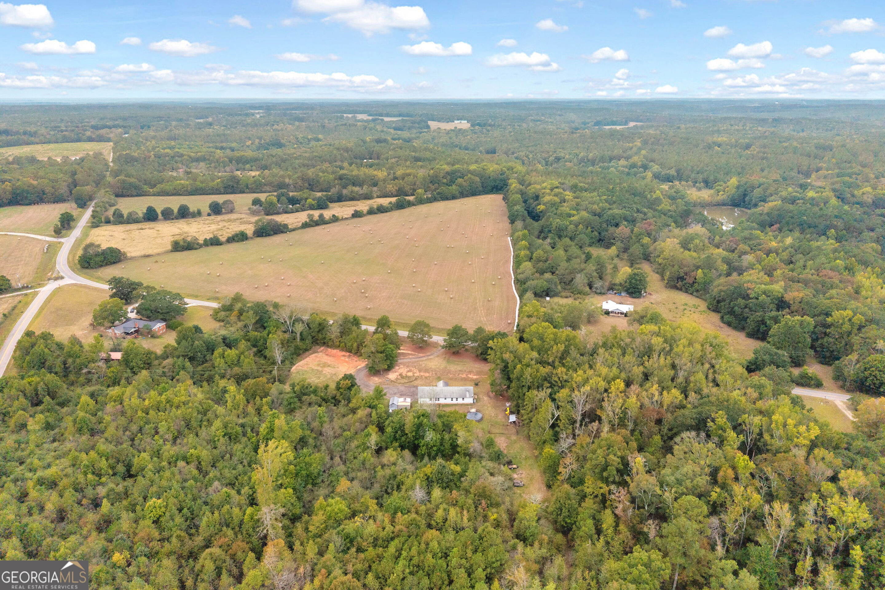 Aerial view of wooded acreage with a cleared homesite, hay fields, and scattered buildings.