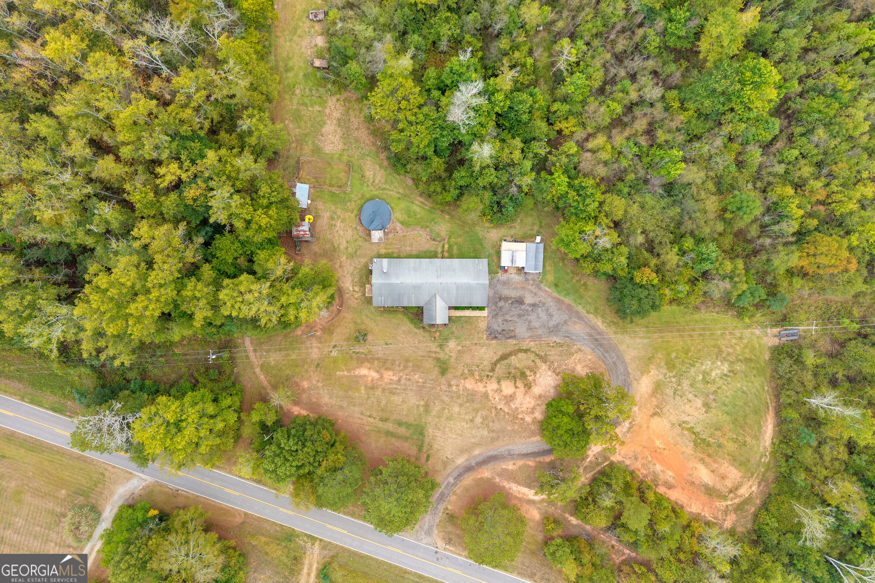 Aerial view of a house in a wooded clearing, with outbuildings, a trampoline, and a driveway to the road.