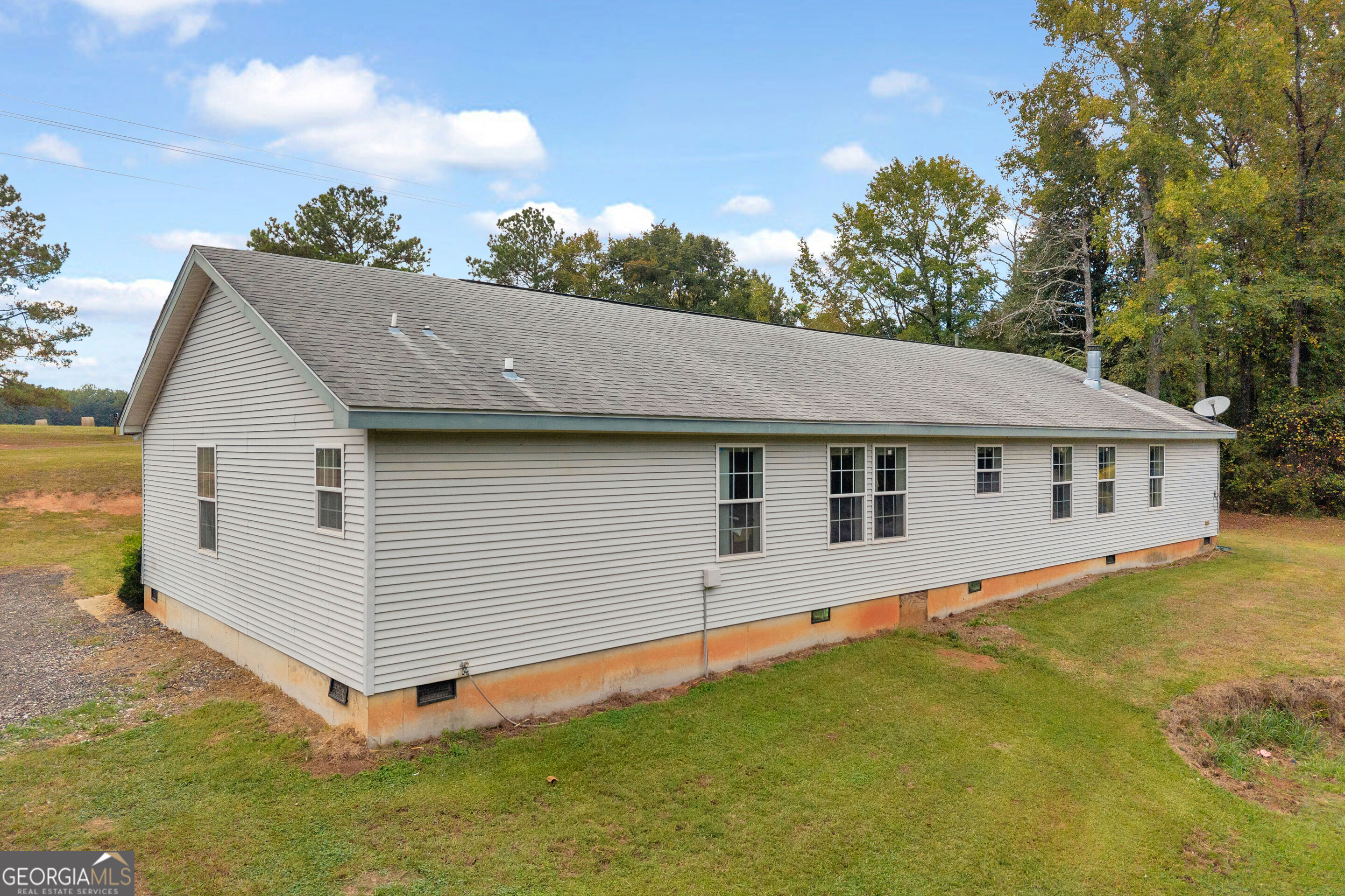 Long, single-story light-sided house with a gray roof, grassy yard, and mature trees.
