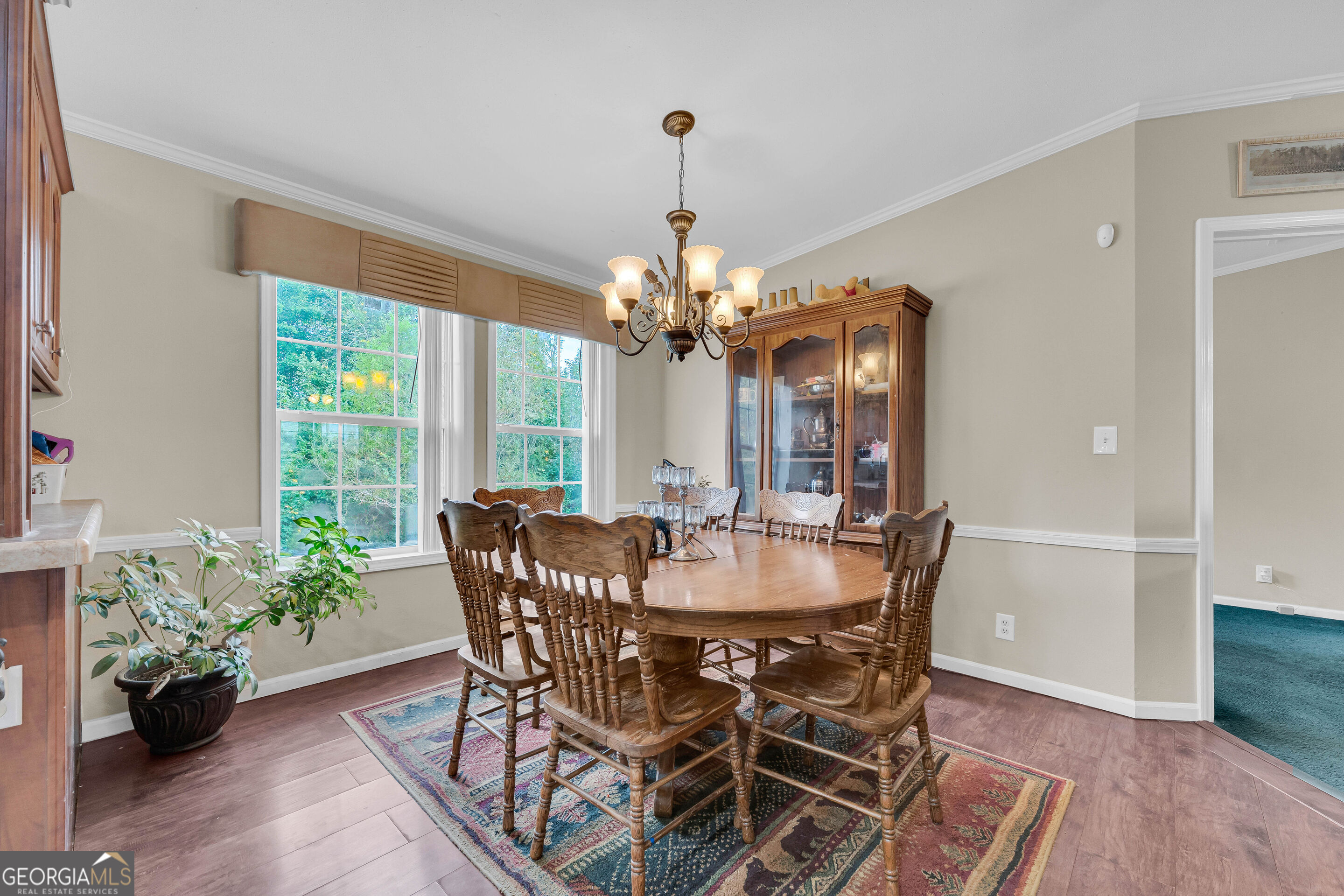 Dining room with a wooden table, glass-front china cabinet, and tall windows overlooking greenery.