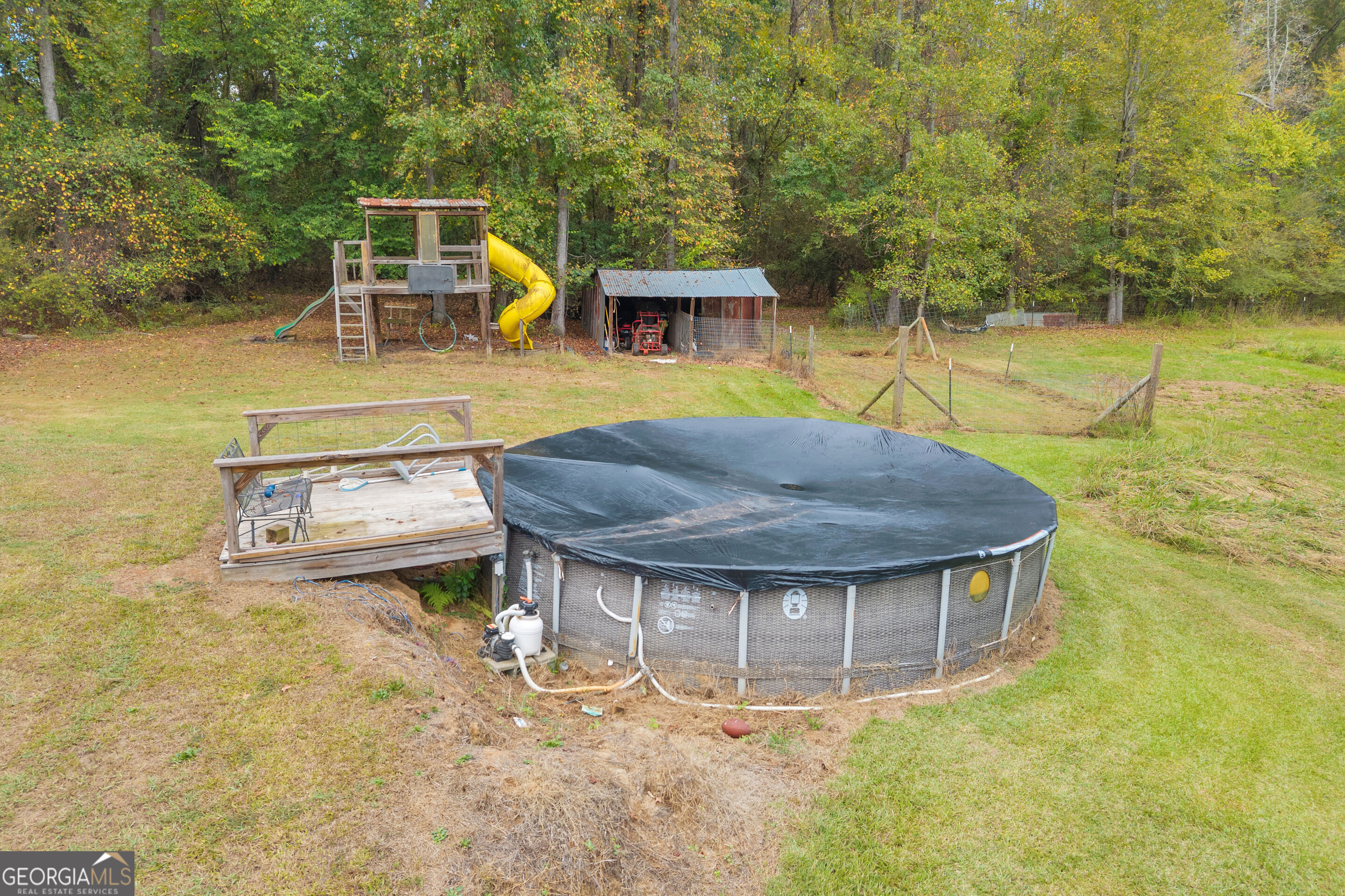 Covered above-ground pool beside a wooden deck and children's playset in a wooded backyard.