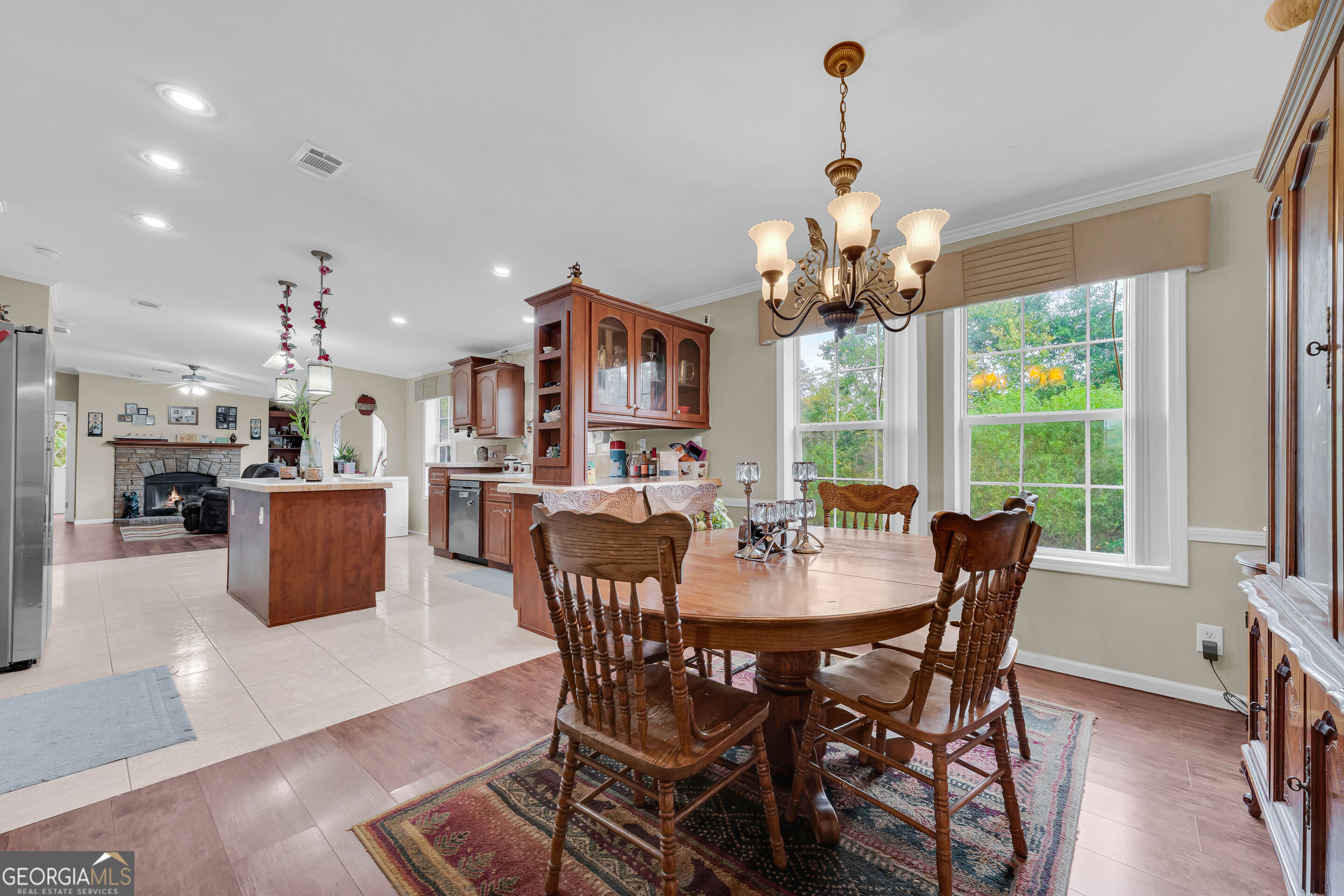 Dining area with a large wooden table, bright windows, and an adjoining kitchen and stone fireplace.