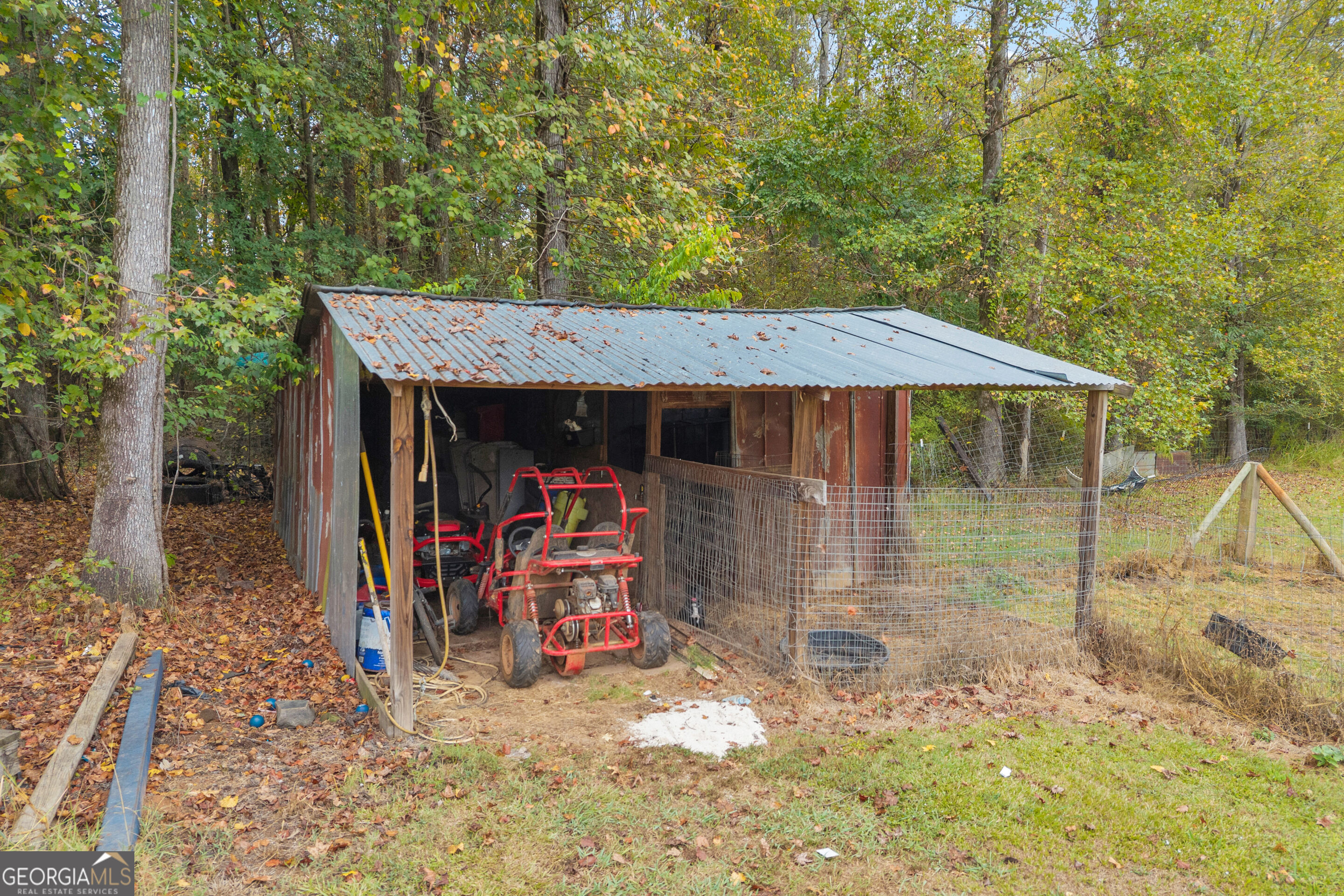 Wooded property outbuilding with a metal roof, fenced enclosure, and red utility vehicles.