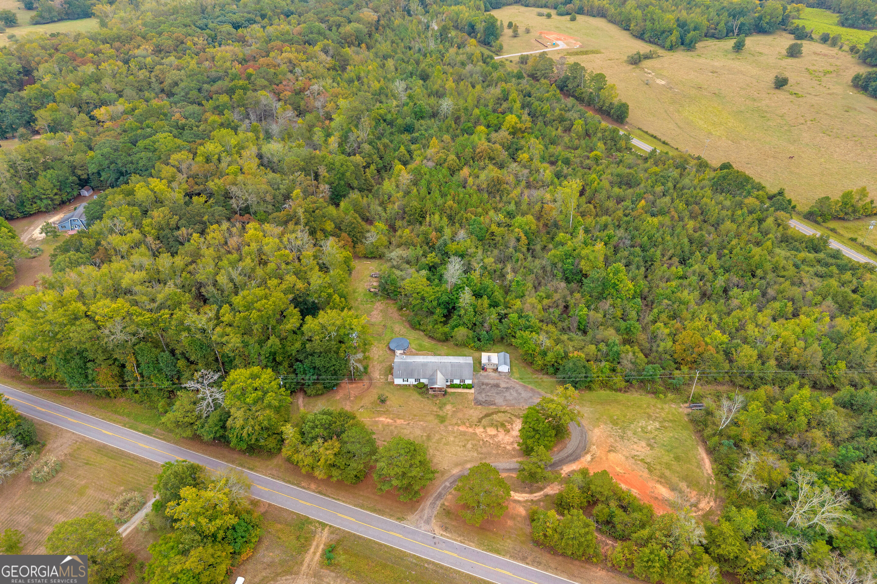 Aerial view of a rural house and outbuildings surrounded by woodland beside a paved road.