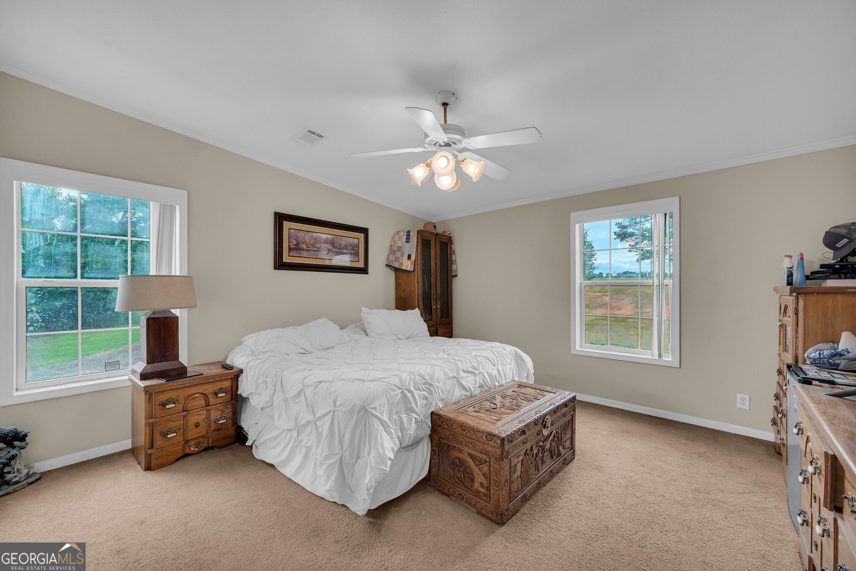 Bedroom with white bedding, two large windows, wooden furniture, and an ornate carved chest.