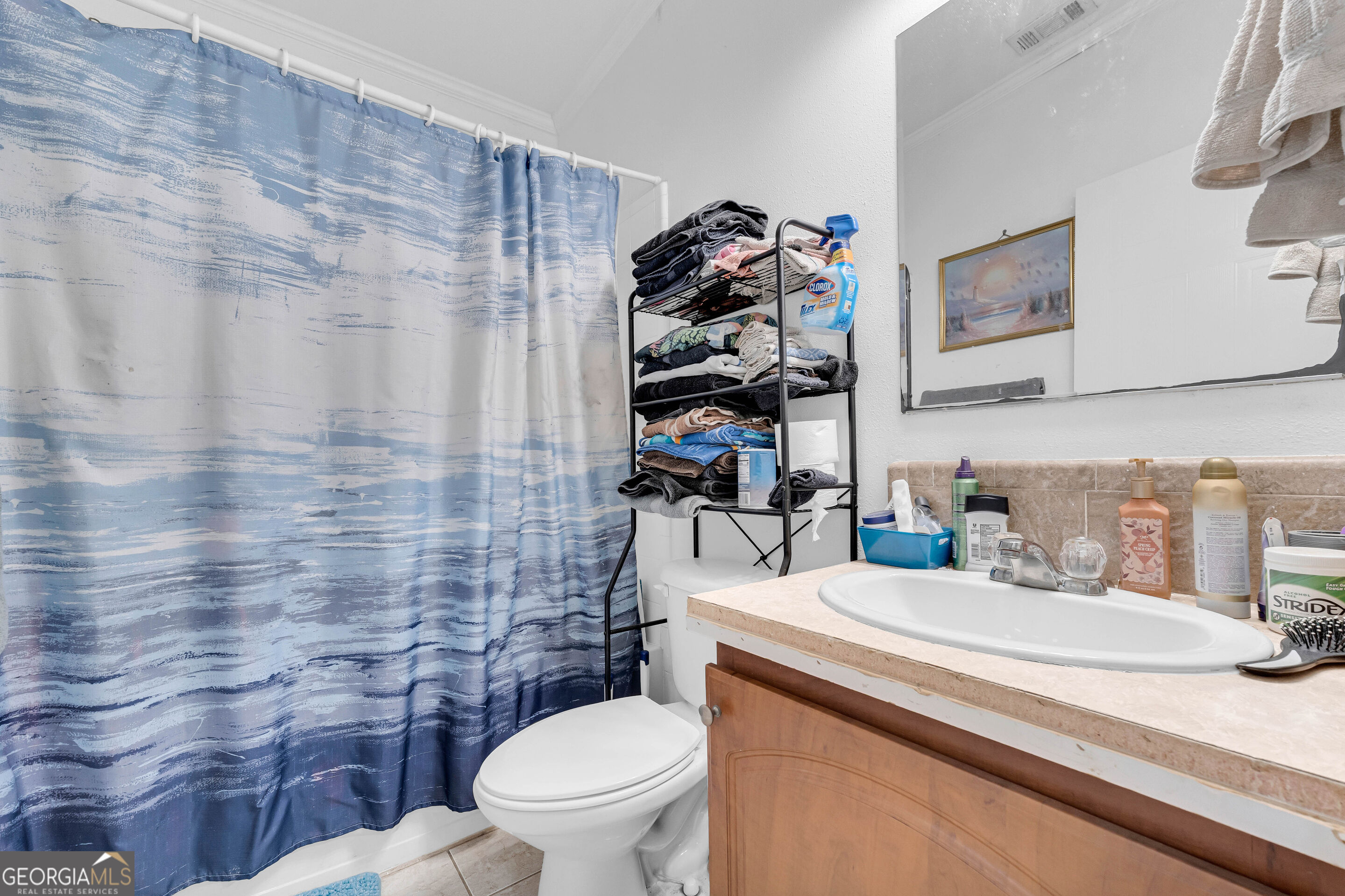 Bathroom with a blue patterned shower curtain, towel-filled shelving rack, and wood vanity.