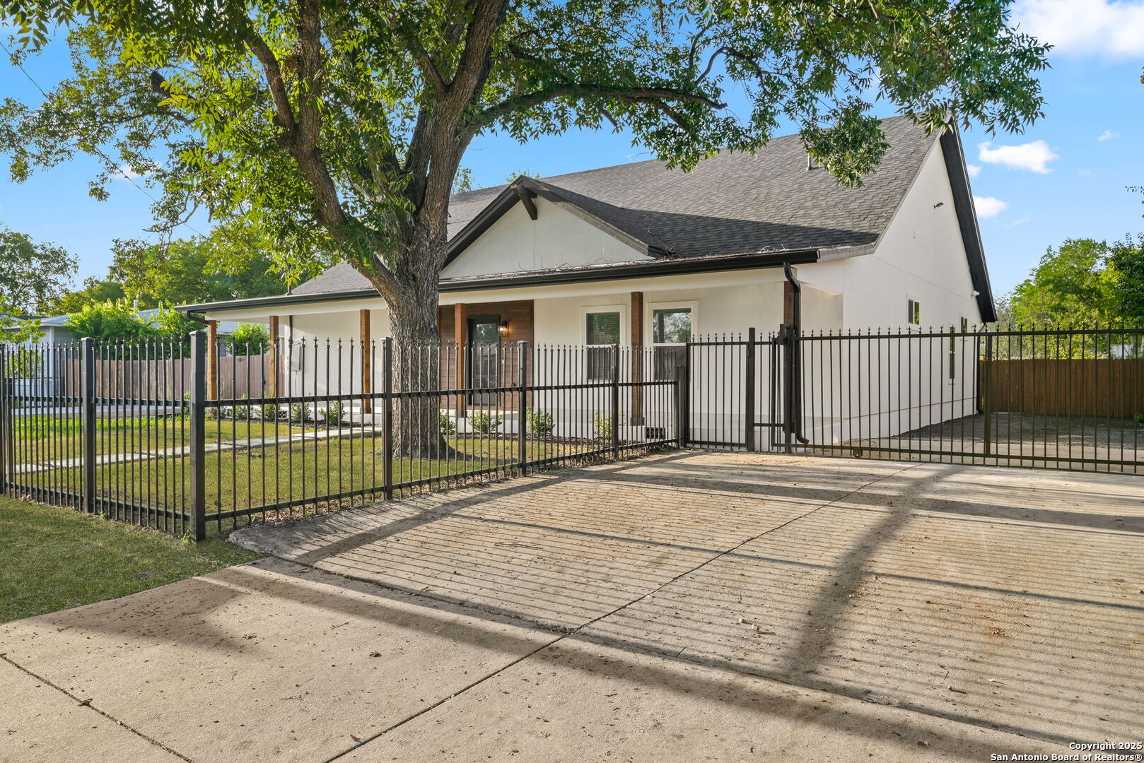 House exterior behind a black metal fence, with a broad concrete driveway and mature tree.