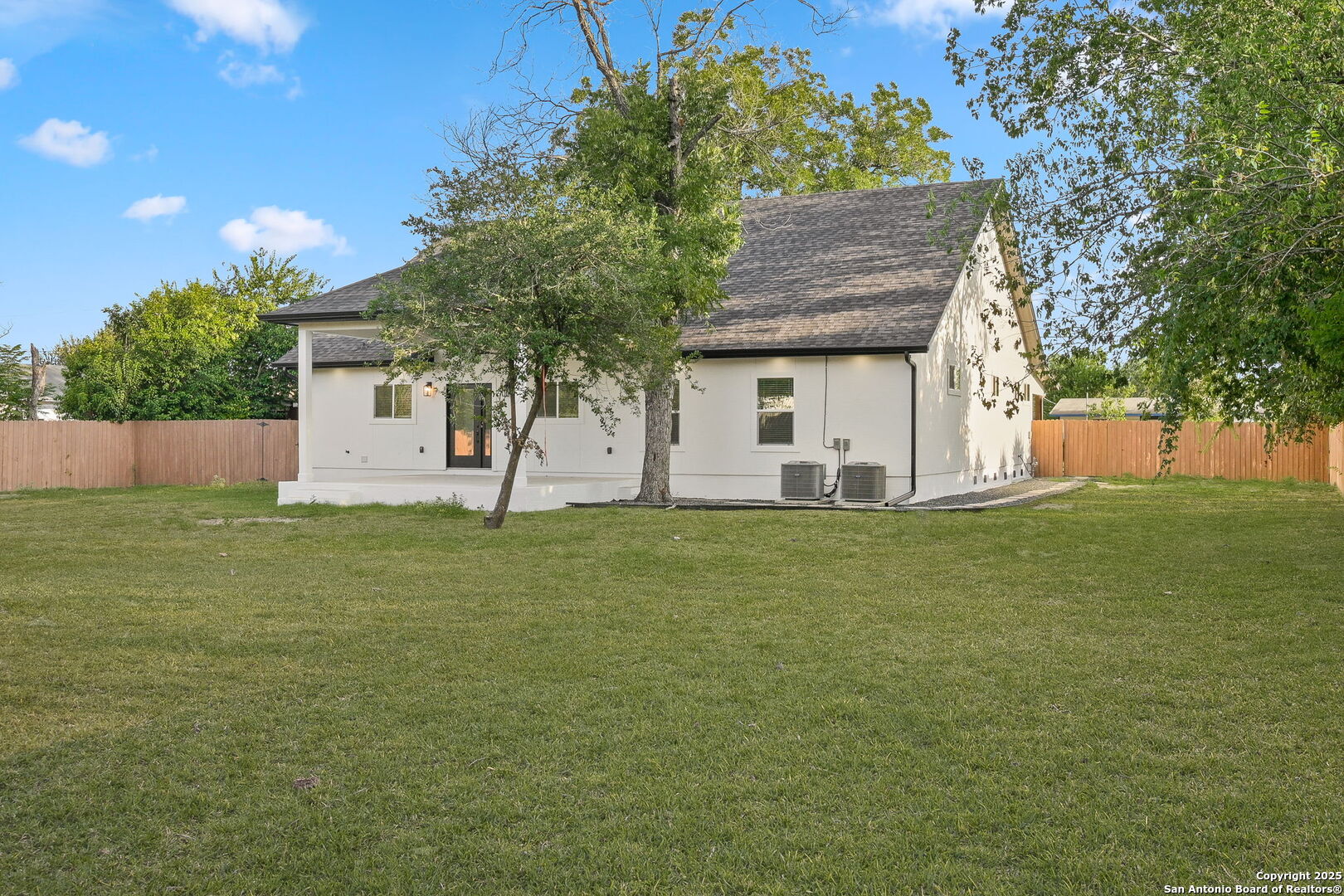 White single-story home's rear exterior with a broad lawn, mature trees, and wooden privacy fencing.