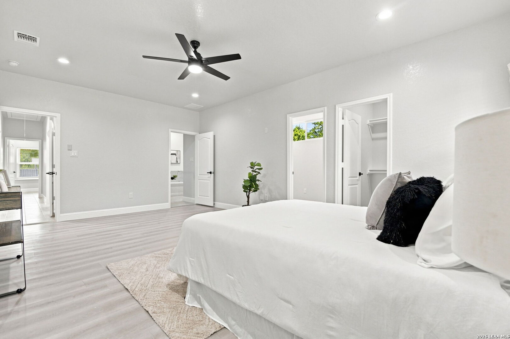 Bright staged bedroom with a white bed, black ceiling fan, and light wood-look flooring.