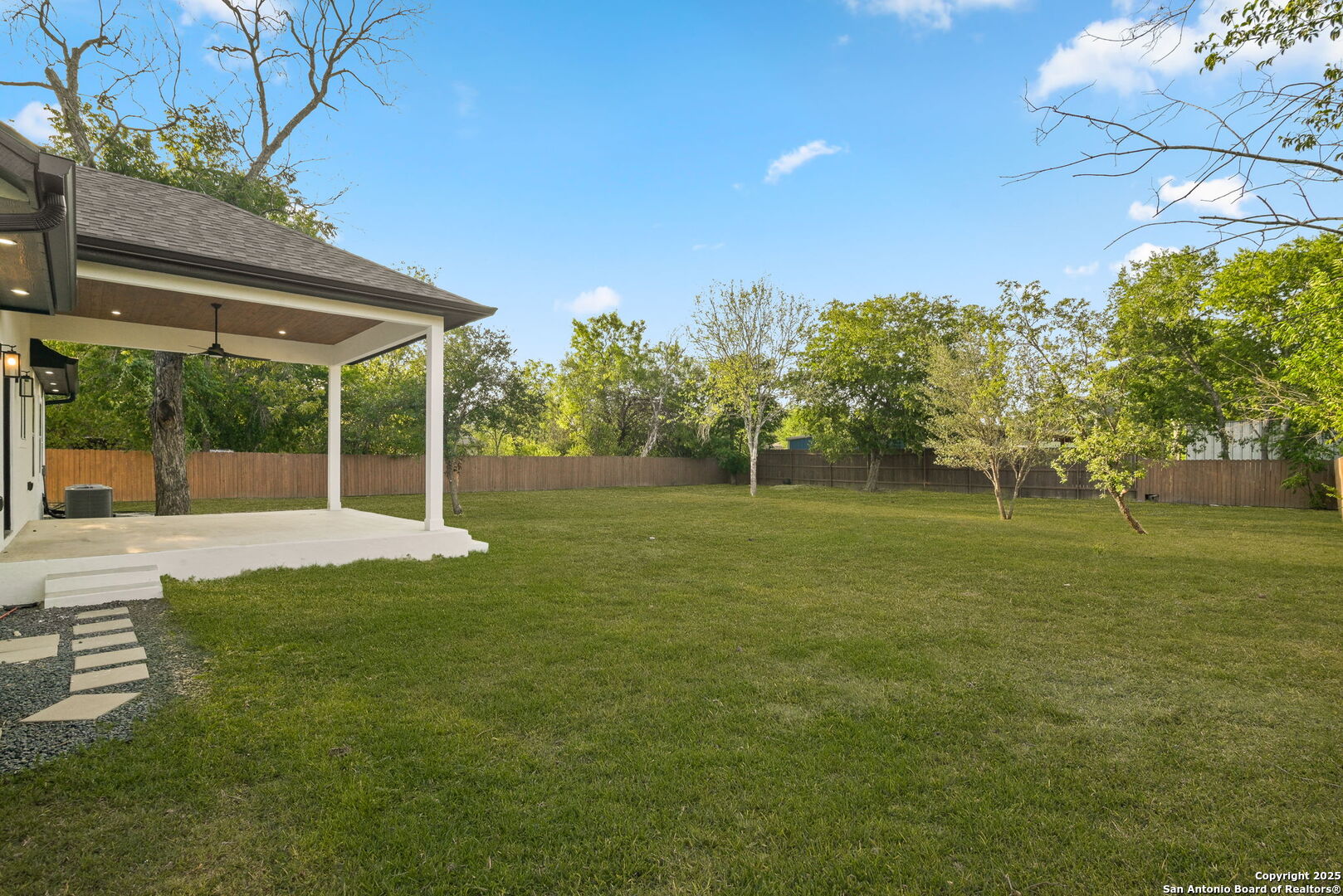 Backyard with a covered patio, broad lawn, mature trees, and a wooden privacy fence.