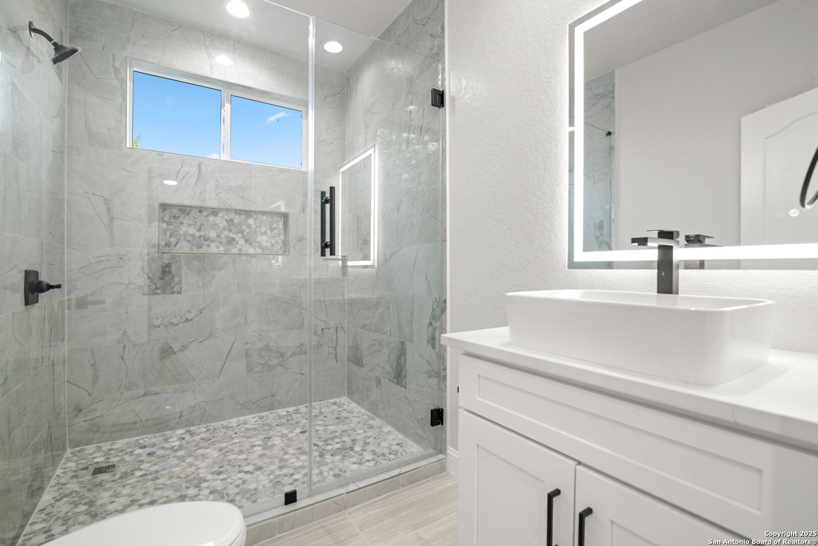 Bathroom with a glass-enclosed marble-look shower, pebble floor, and white vessel sink.