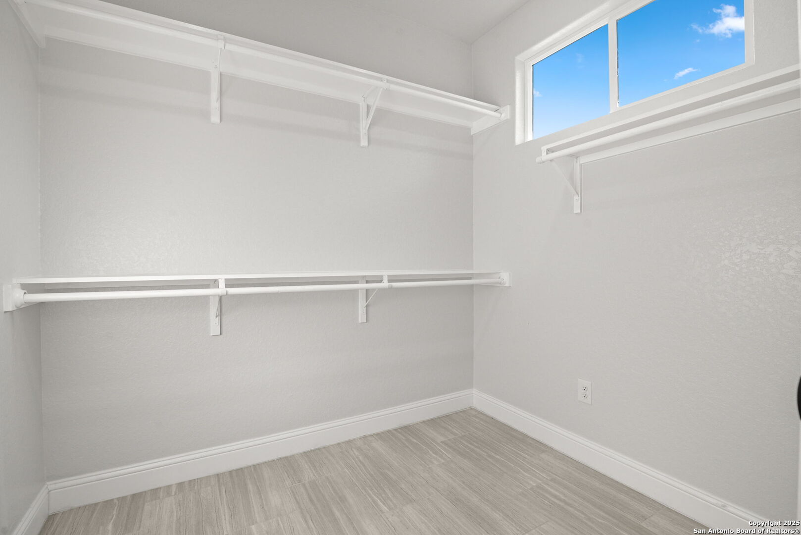 Empty walk-in closet with white shelving and hanging rods, gray plank flooring, and a high window.