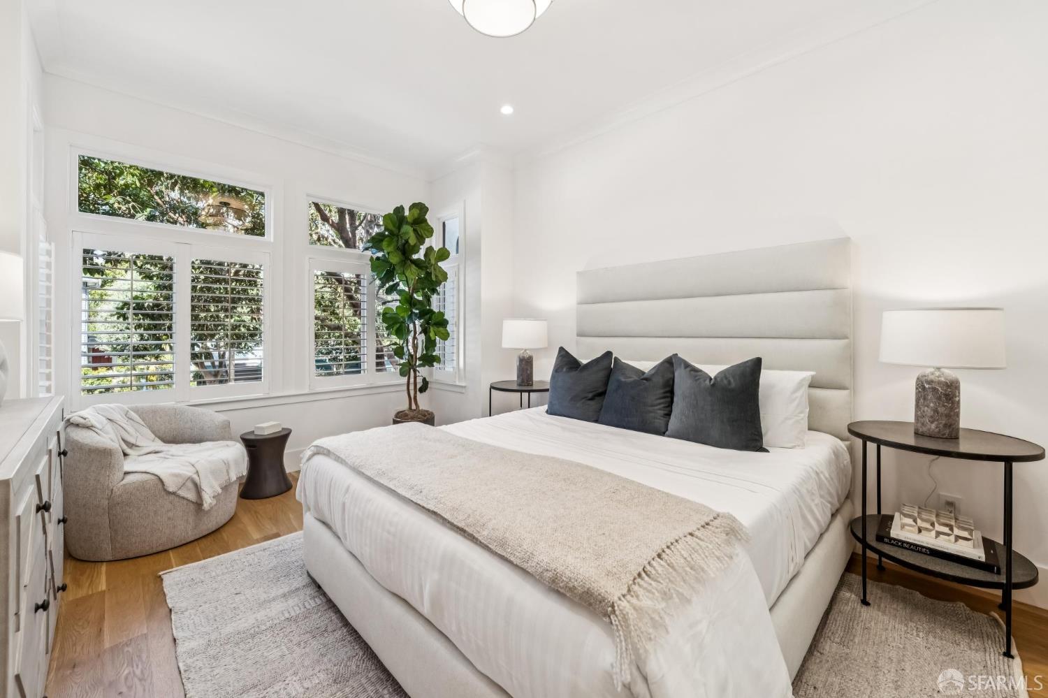 Staged bedroom with a large upholstered bed, shuttered windows, and a cozy reading chair.