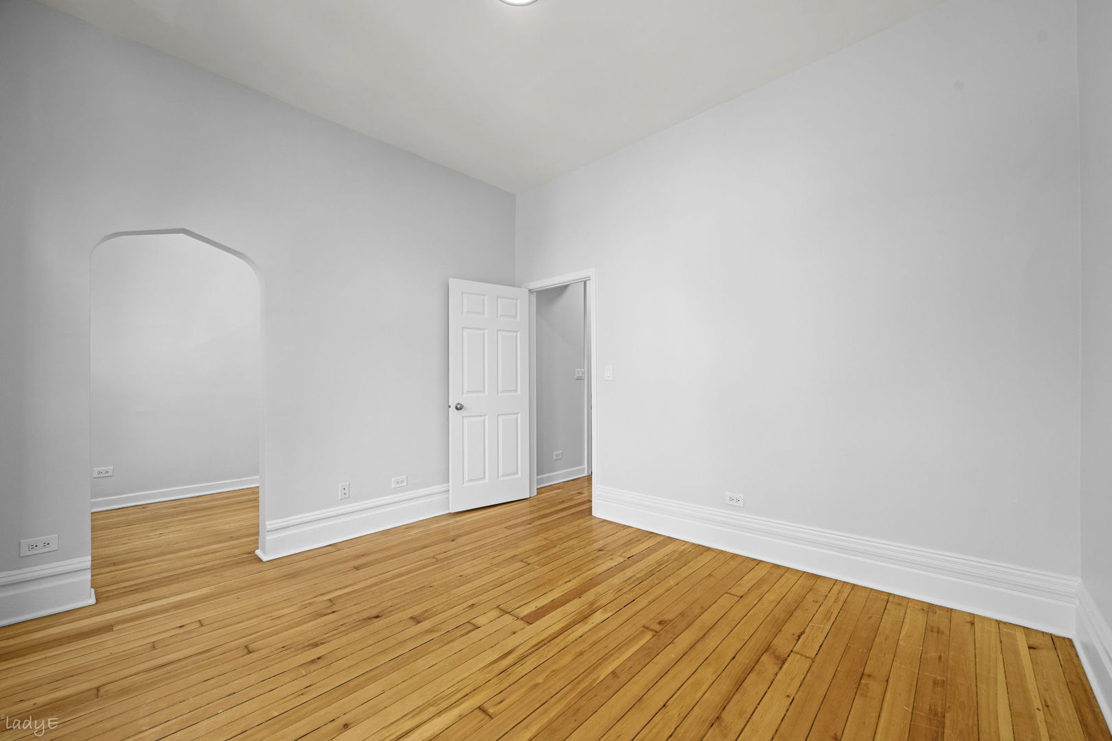 Vacant room with honey-colored hardwood floors, pale walls, an arched opening, and an open door.