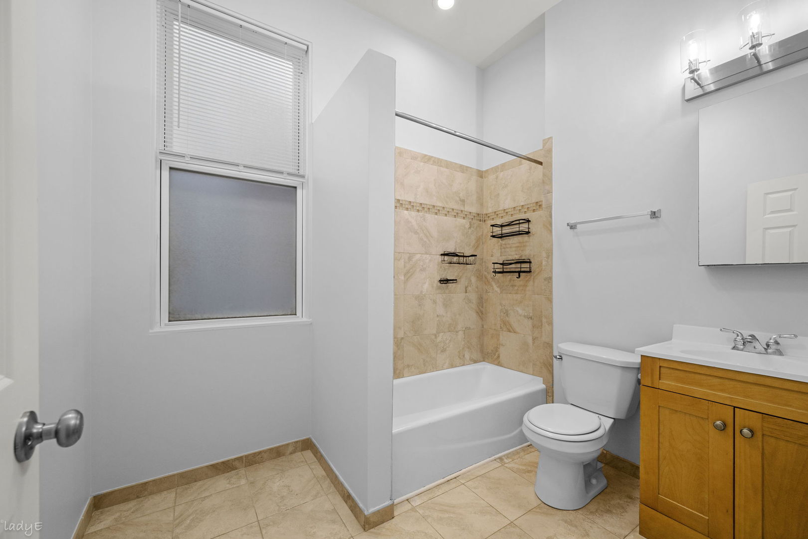 Bathroom with beige tiled tub surround, window with blinds, toilet, and wood vanity.