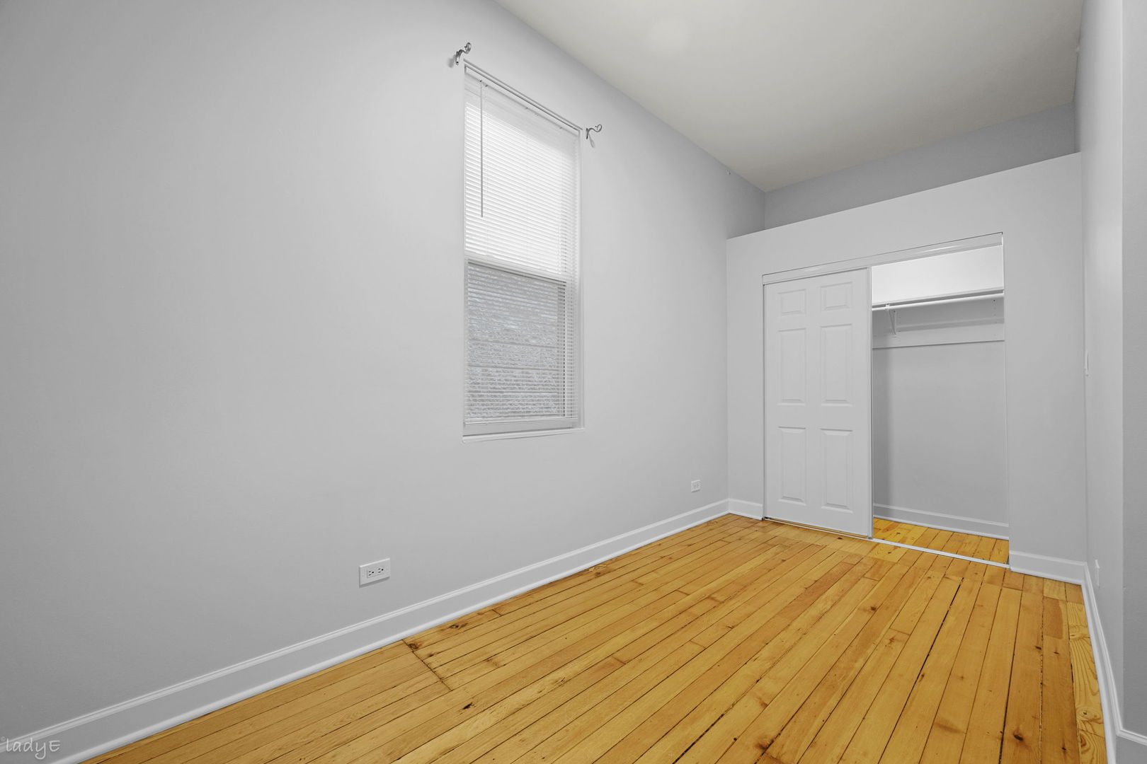 Empty bedroom with hardwood floors, a tall window, and a partially open closet.