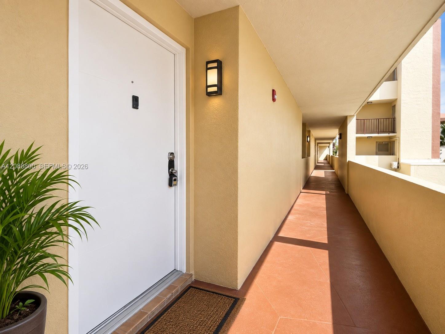 Condominium entrance along a sunlit covered corridor, with a white door and terracotta tile walkway.