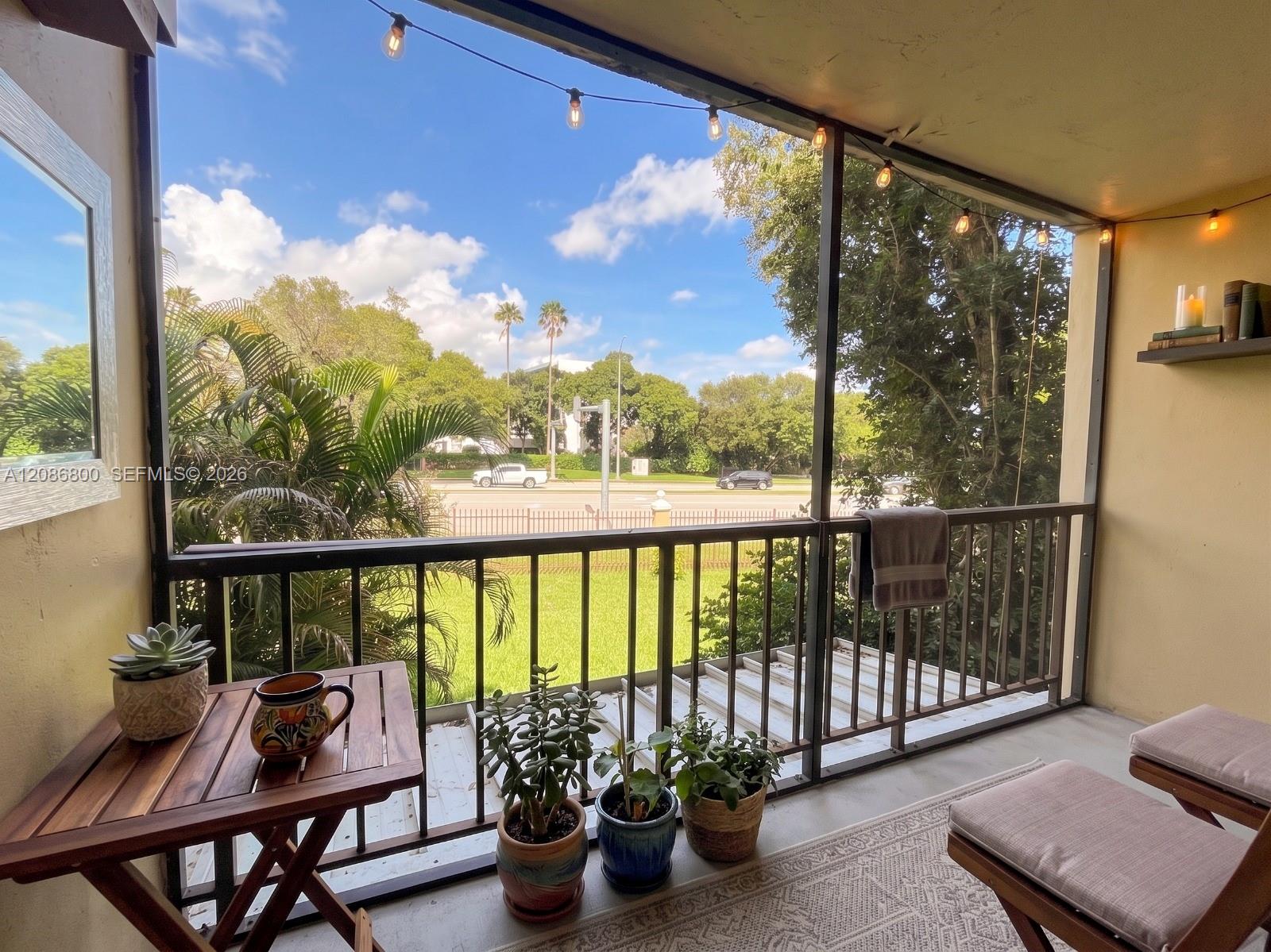Furnished balcony with potted plants and string lights overlooking trees and a street.
