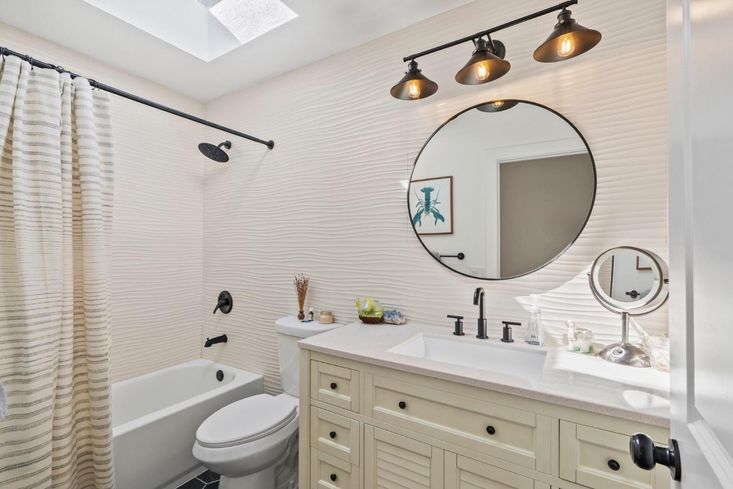 Bathroom with a pale vanity, large round mirror, wavy white tile, and bathtub behind a striped curtain.