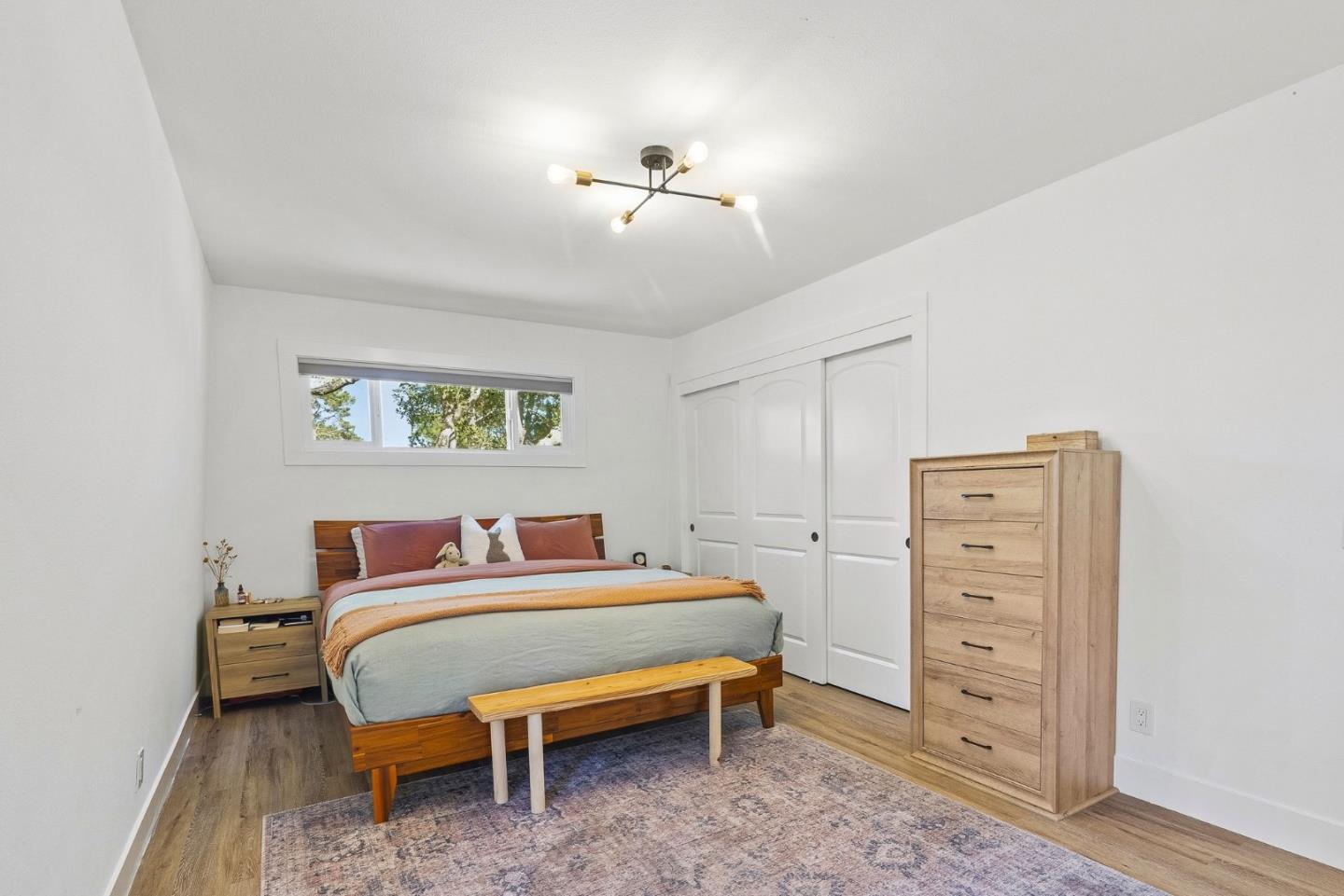 Staged bedroom with a wooden bed, tall dresser, sliding closet doors, and a narrow window overlooking trees.