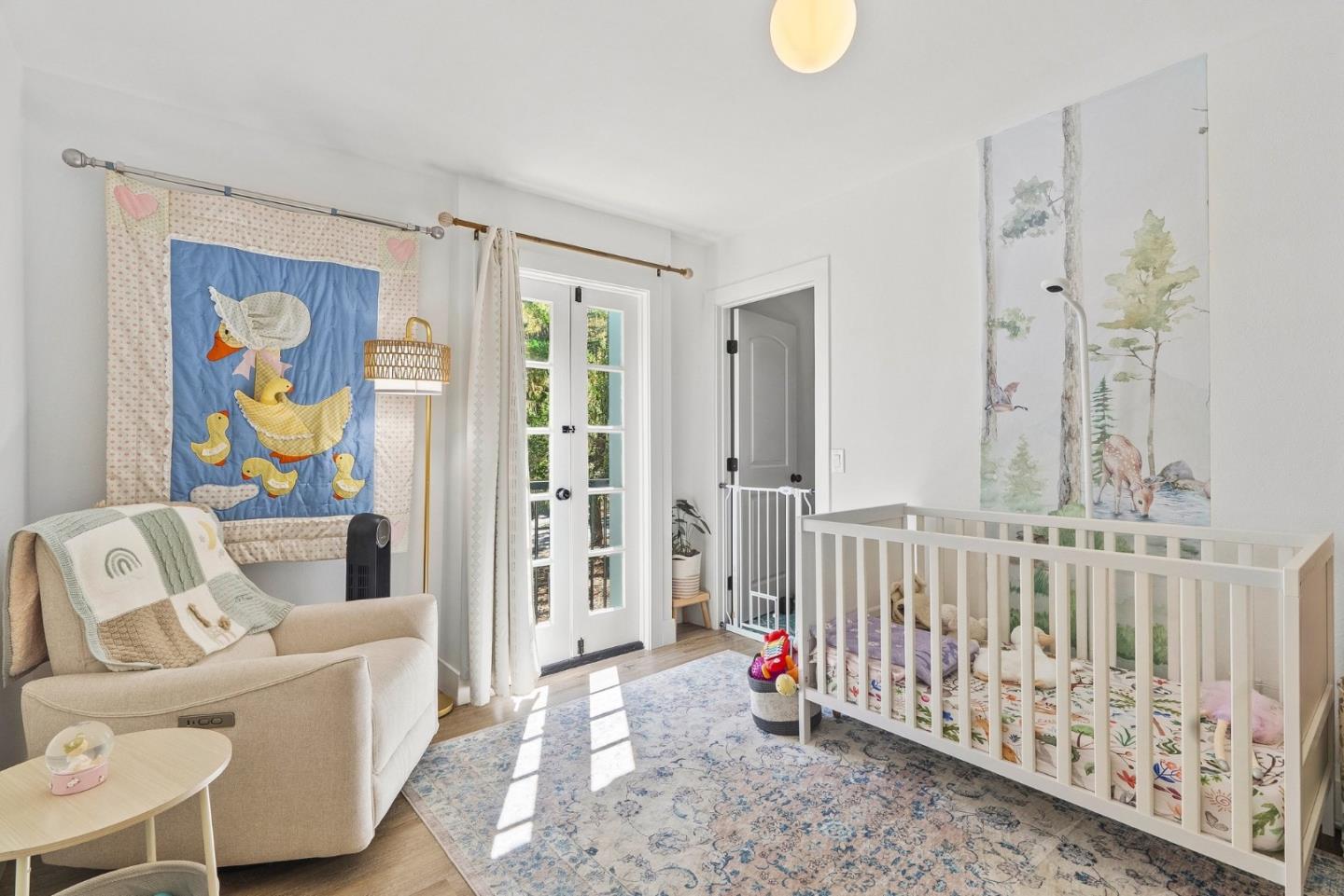 Nursery with a white crib, woodland mural, and glider beside French doors.