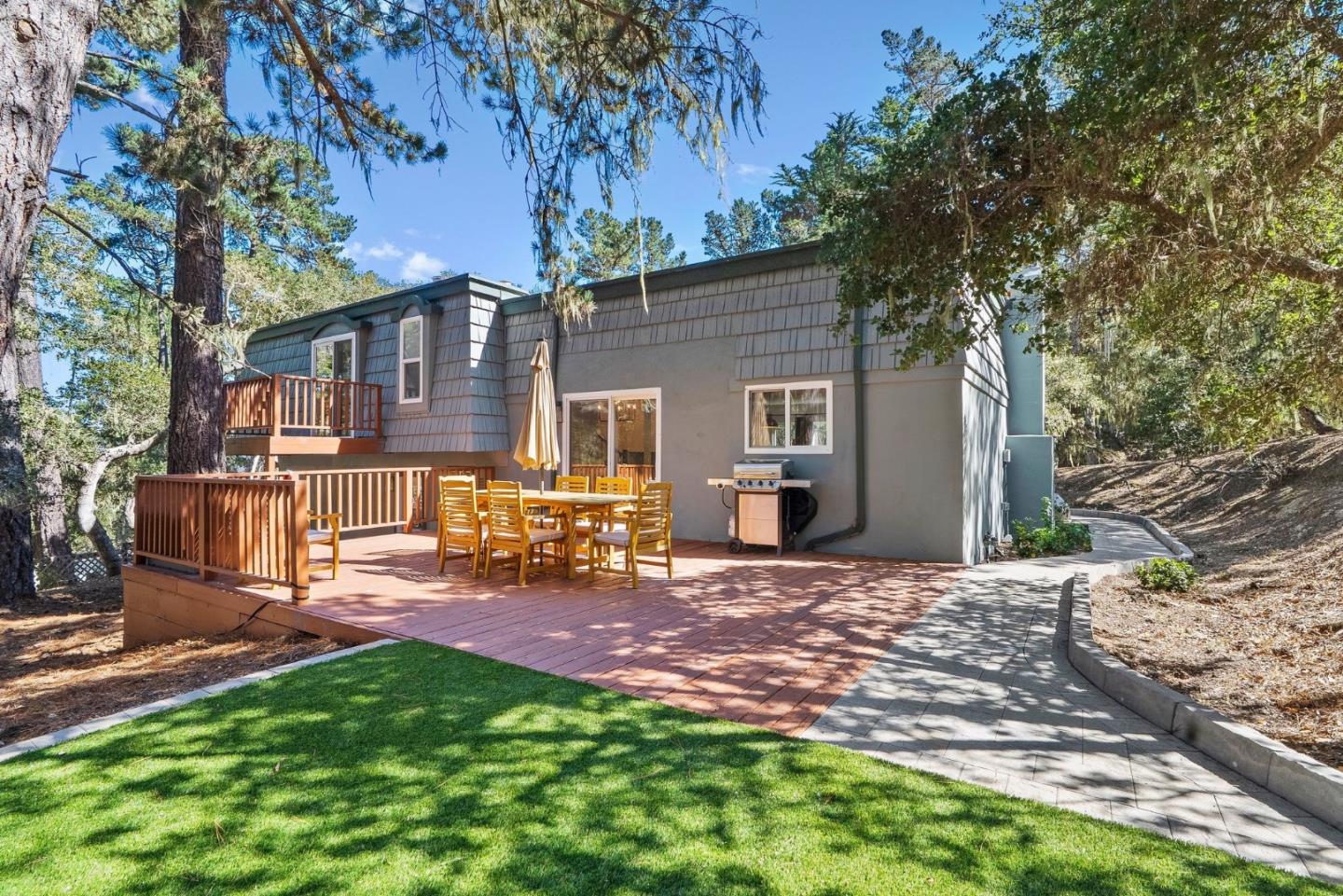 Gray Monterey home with a broad deck, outdoor dining set, and grill among mature trees.