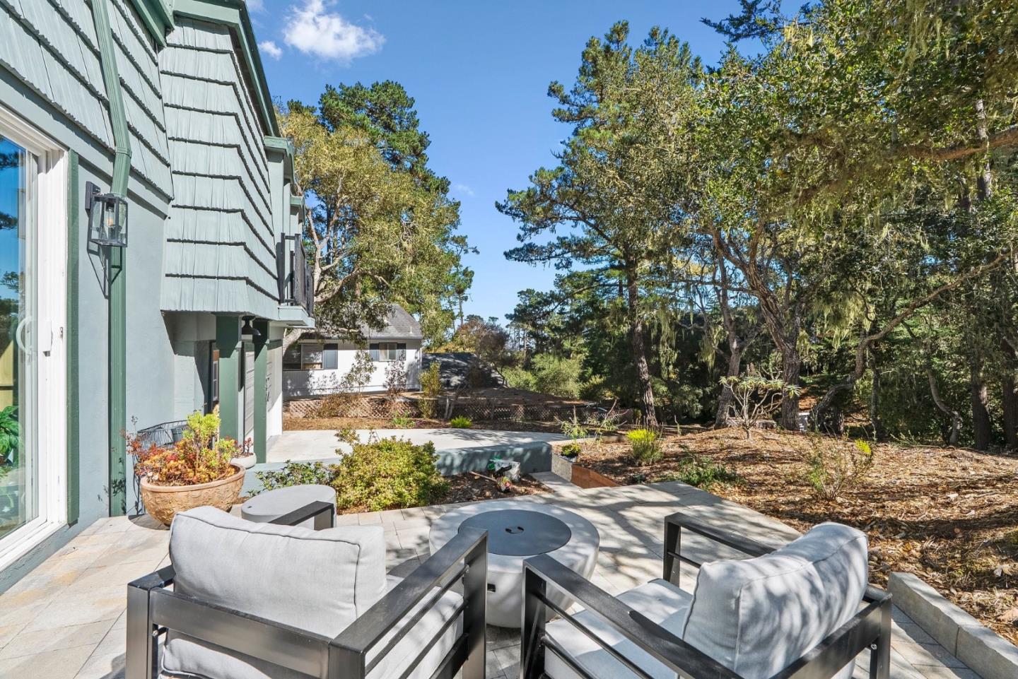 Patio beside a pale green house with cushioned seating, a round table, and mature trees.