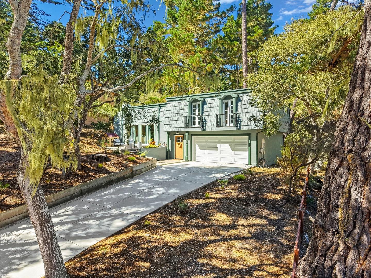 Two-story gray-green house with a yellow front door, white garage, and tree-lined driveway.