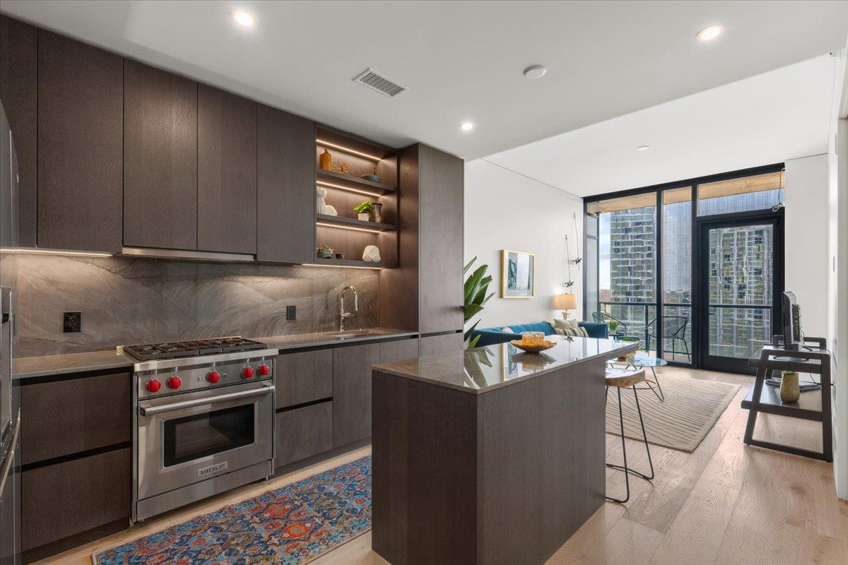 Open-plan condo kitchen with dark cabinetry, marble backsplash, and island beside a windowed living area.