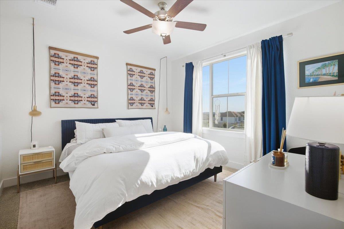 Furnished bedroom with a white bed, dark blue headboard, and blue curtains around a sunny window.
