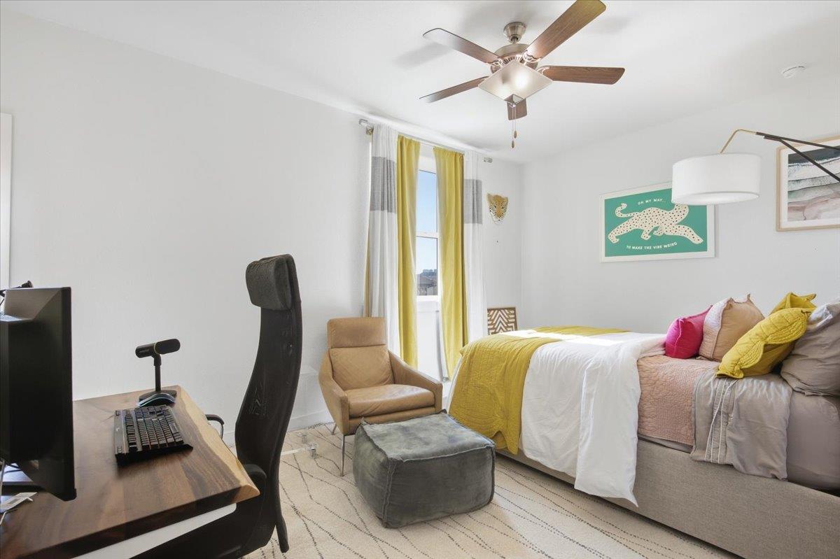 Bedroom with a mustard-yellow bedspread, ceiling fan, and a desk beside a sitting area.