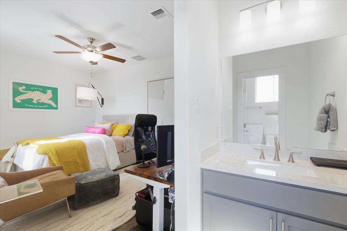Furnished bedroom beside a bathroom vanity, with yellow bedding, colorful pillows, and a large mirror.