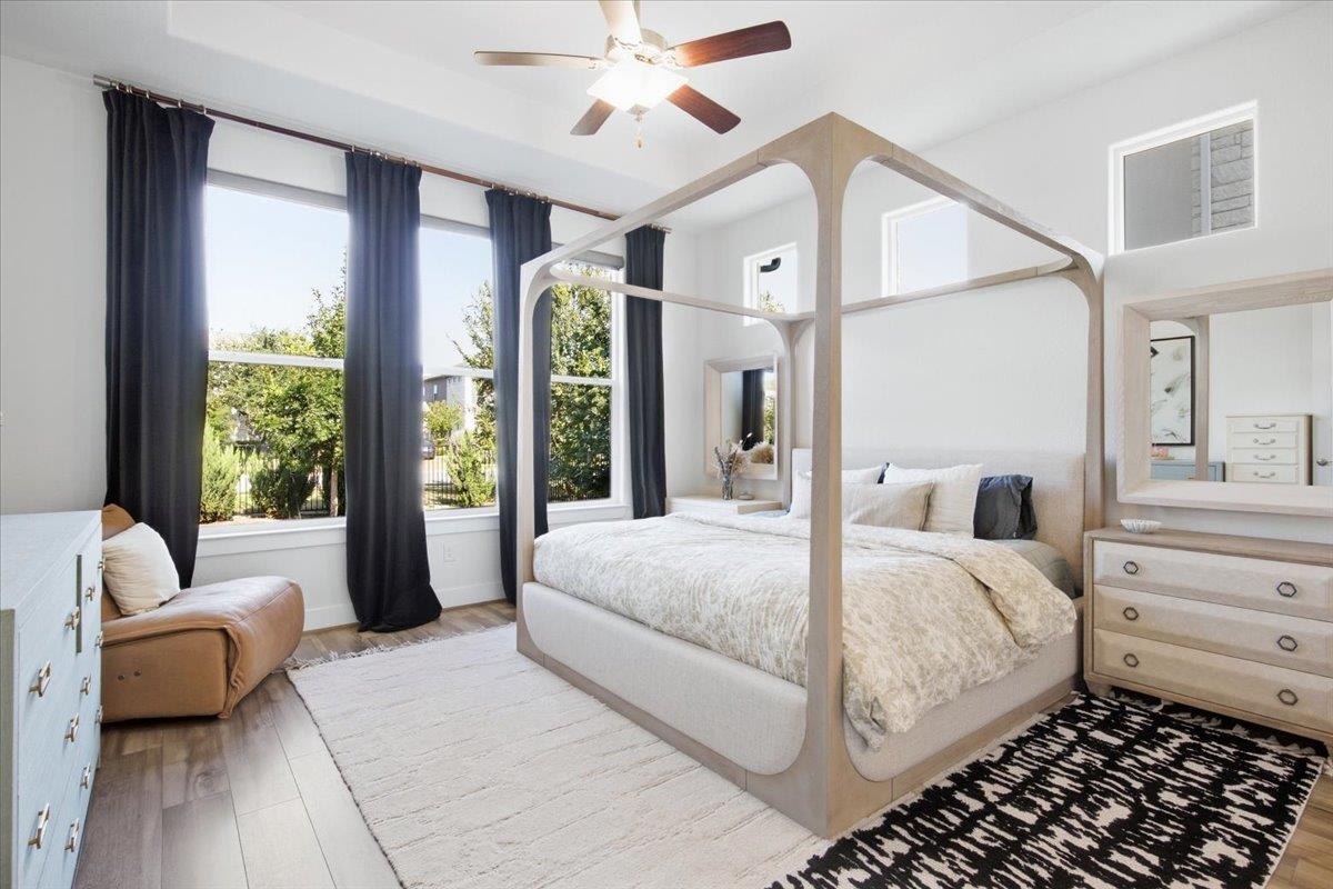 Bright bedroom with a light wood canopy bed, tall curtained windows, and a ceiling fan.
