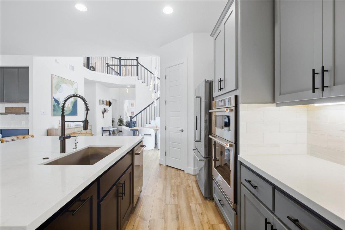 Modern kitchen with a dark island, white counters, stainless steel appliances, and an open staircase beyond.