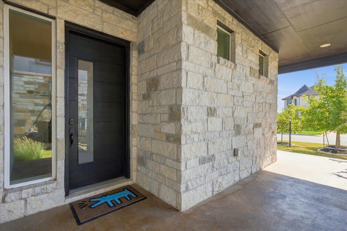 Covered stone entry with a black front door, tall glass panel, and blue WELCOME doormat.