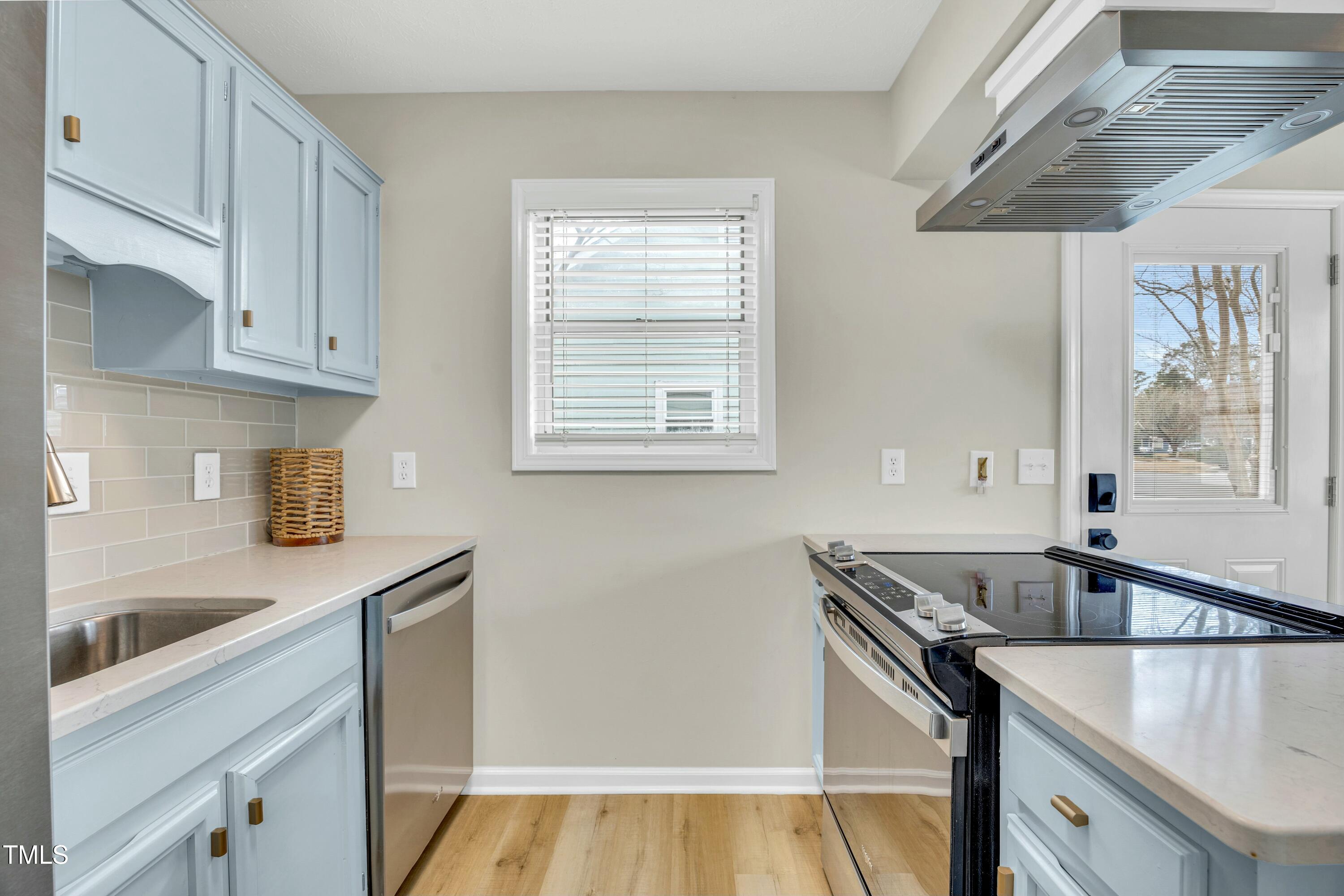 Kitchen with pale blue cabinets, brass hardware, stainless-steel dishwasher, and black glass cooktop.