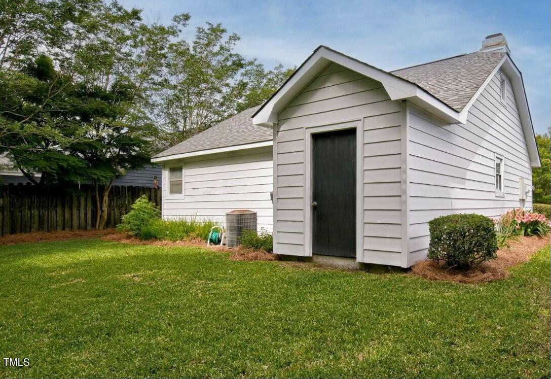 White clapboard outbuilding with a dark door and gray shingle roof beside a green lawn.