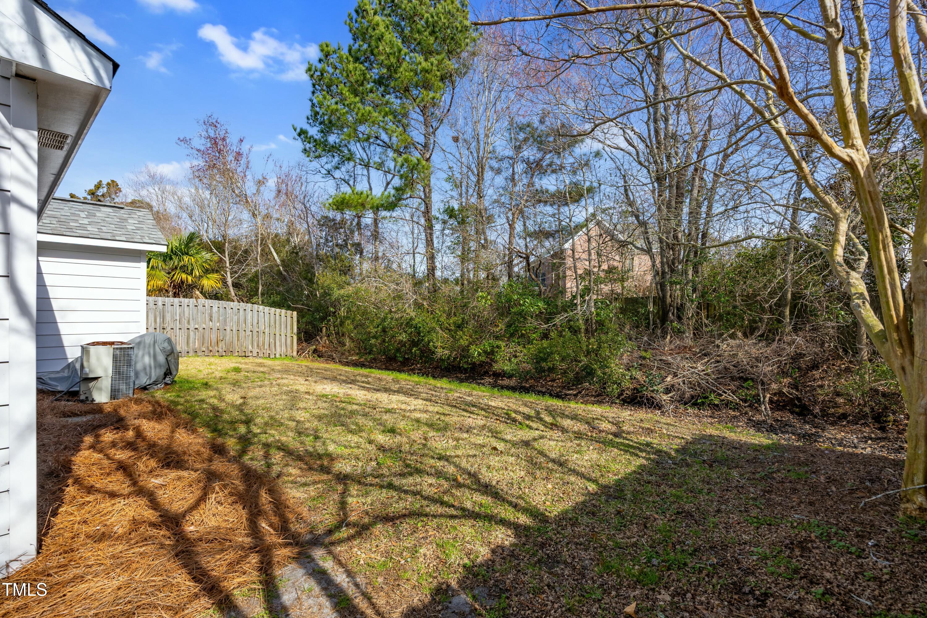 Side yard with a small lawn, wooden fence, and wooded privacy buffer beside a white-sided house.