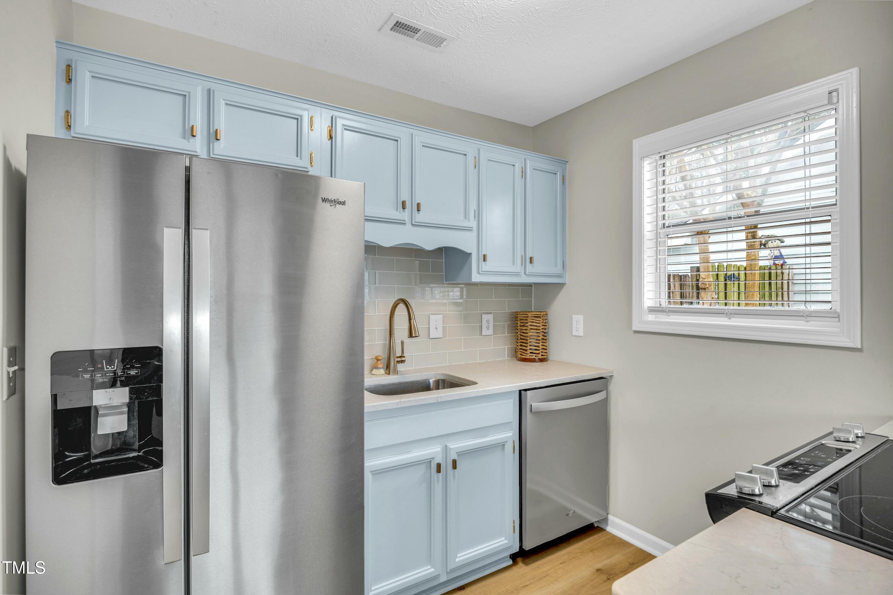 Kitchen with pale blue cabinets, stainless steel appliances, and a window overlooking a fenced yard.