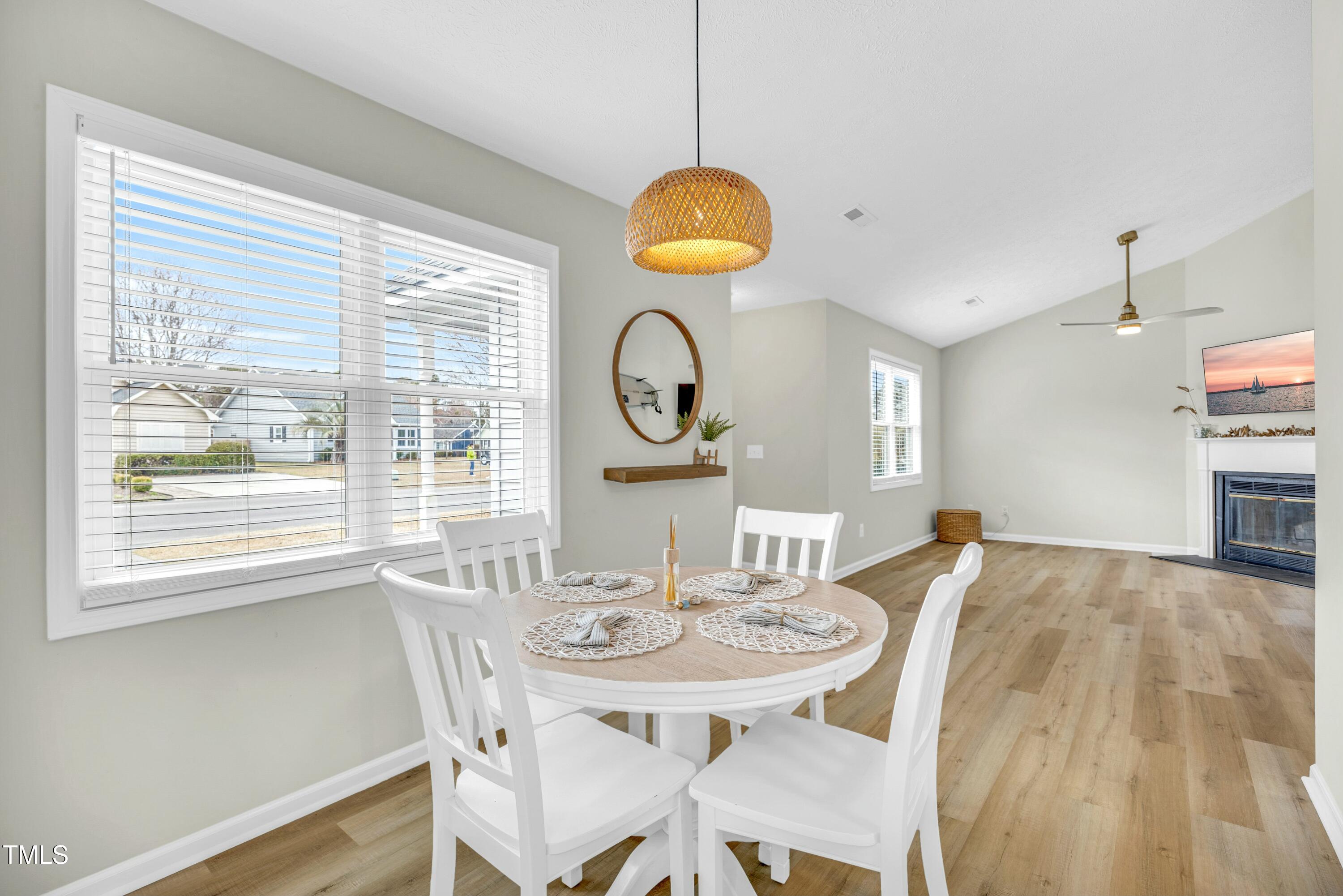 Dining area with a round white table beneath a woven pendant, opening onto a living room with a fireplace.