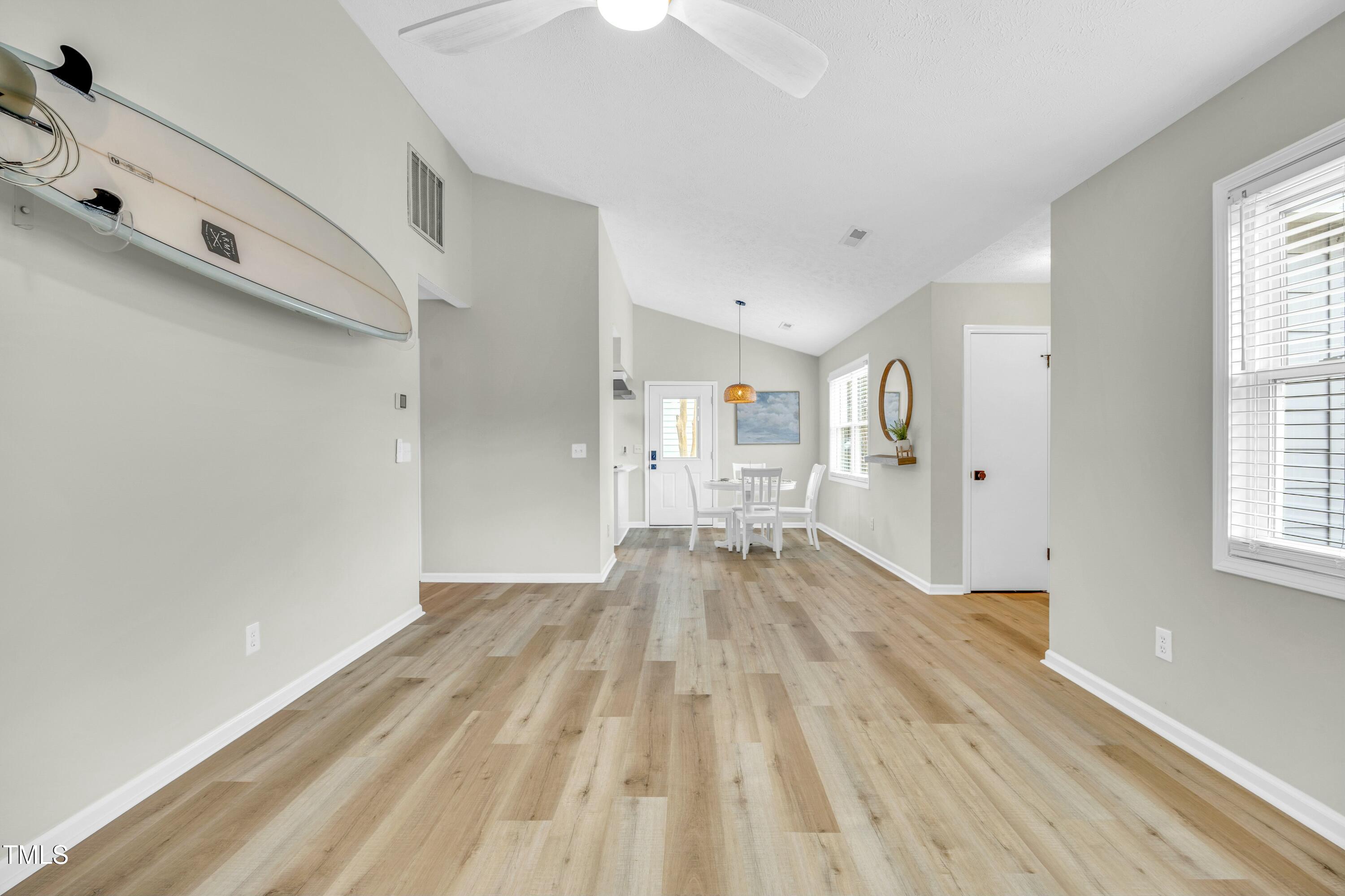 Open dining area with light wood-look floors, white table and chairs, and surfboards mounted on the wall.